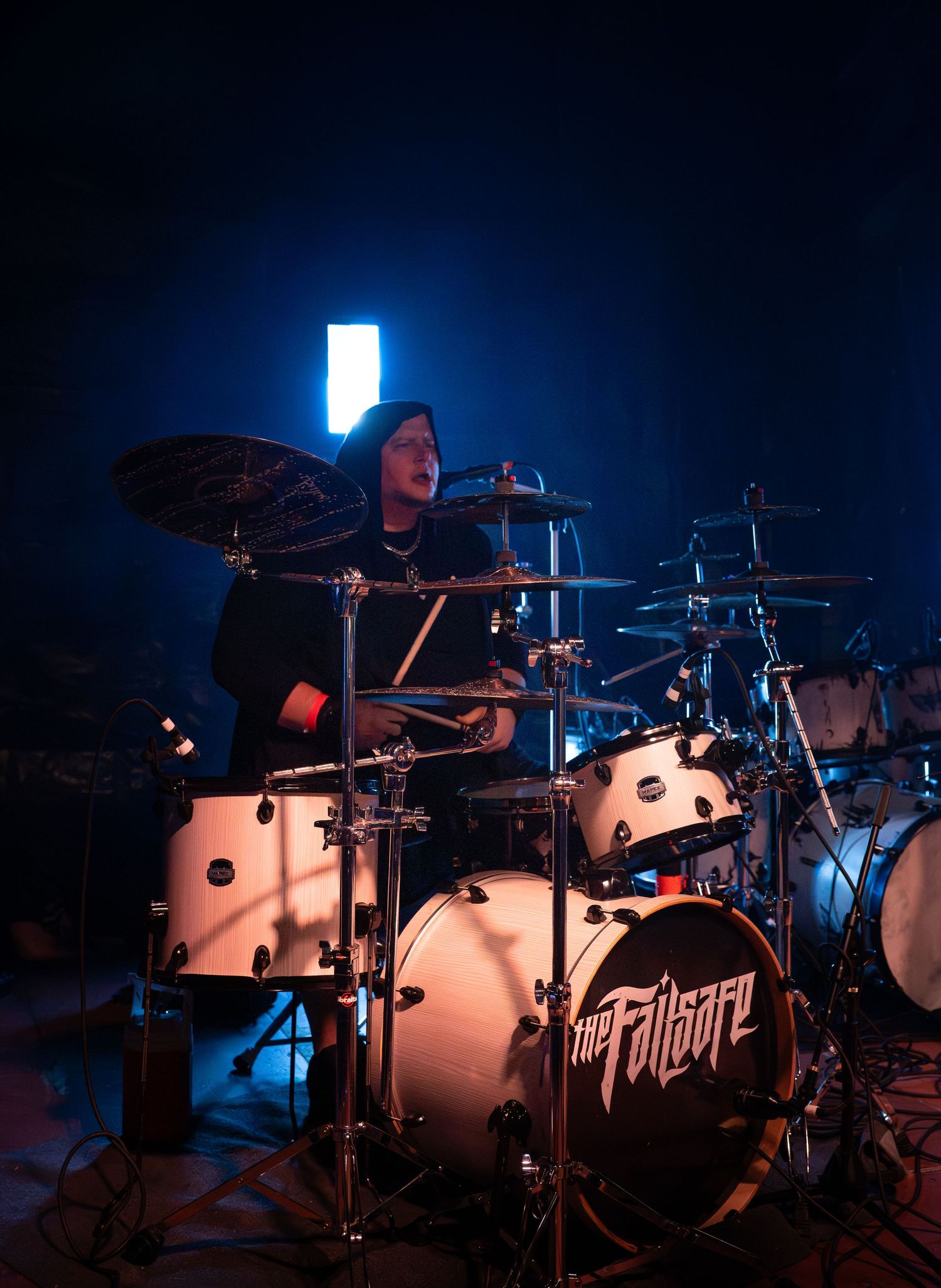 Drummer performs on white polka dot drum kit on dark stage with dramatic blue backlighting during concert.