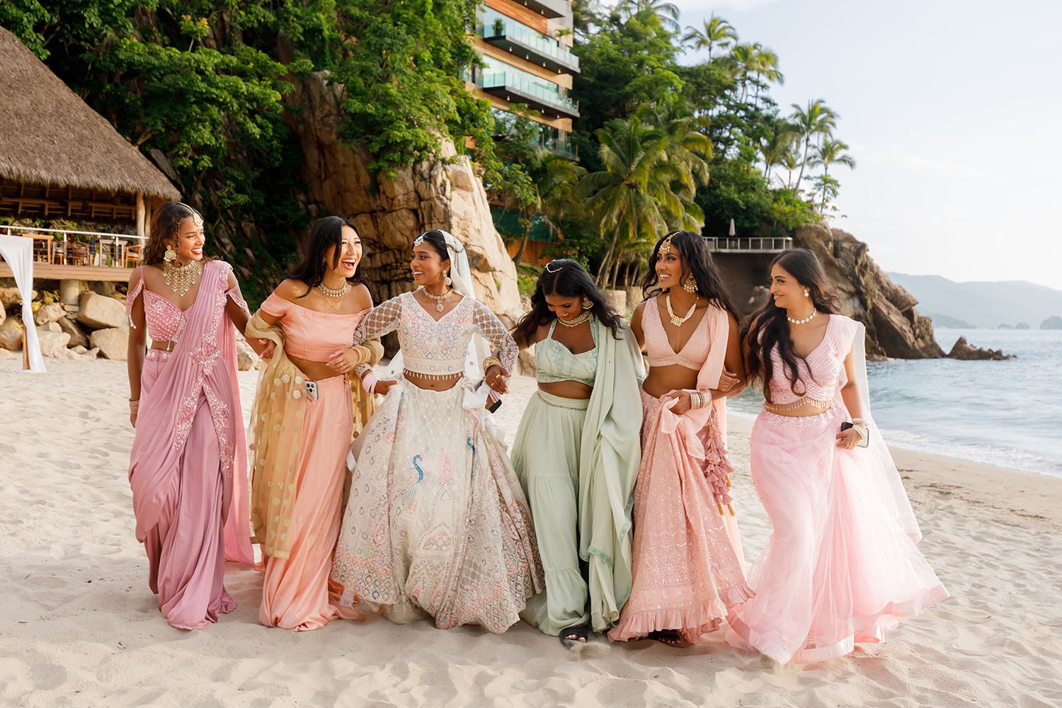 Bridesmaids in pastel lehengas laughing with the bride on the beach during an Indian wedding in Puerto Vallarta, by José Carrillo.