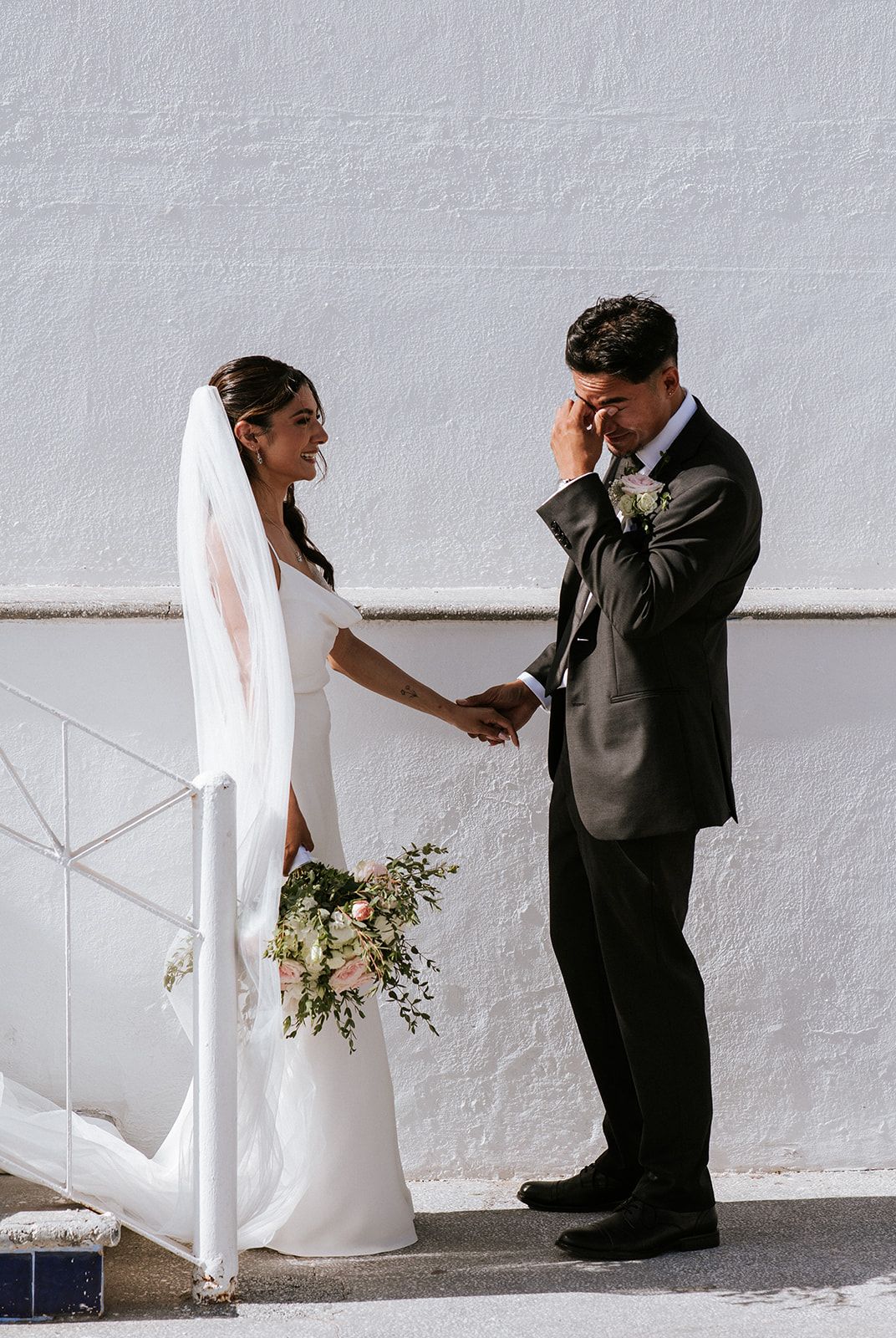 Bride smiles as emotional groom wipes his tears during their first look by a white wall in Puerto Vallarta, by José Carrillo.