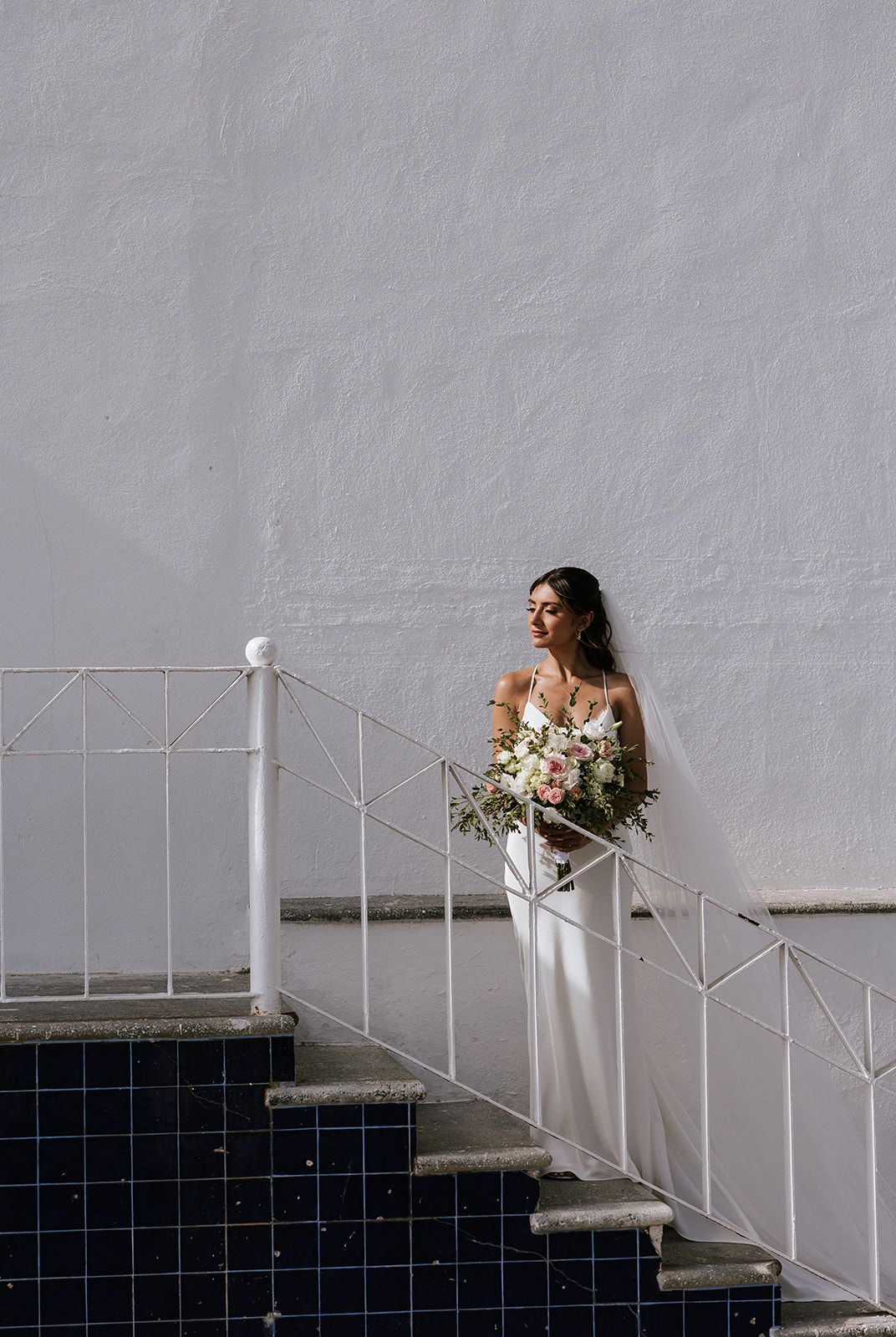Bride standing on white staircase holding pastel bouquet during her wedding in Puerto Vallarta, by José Carrillo Photography.