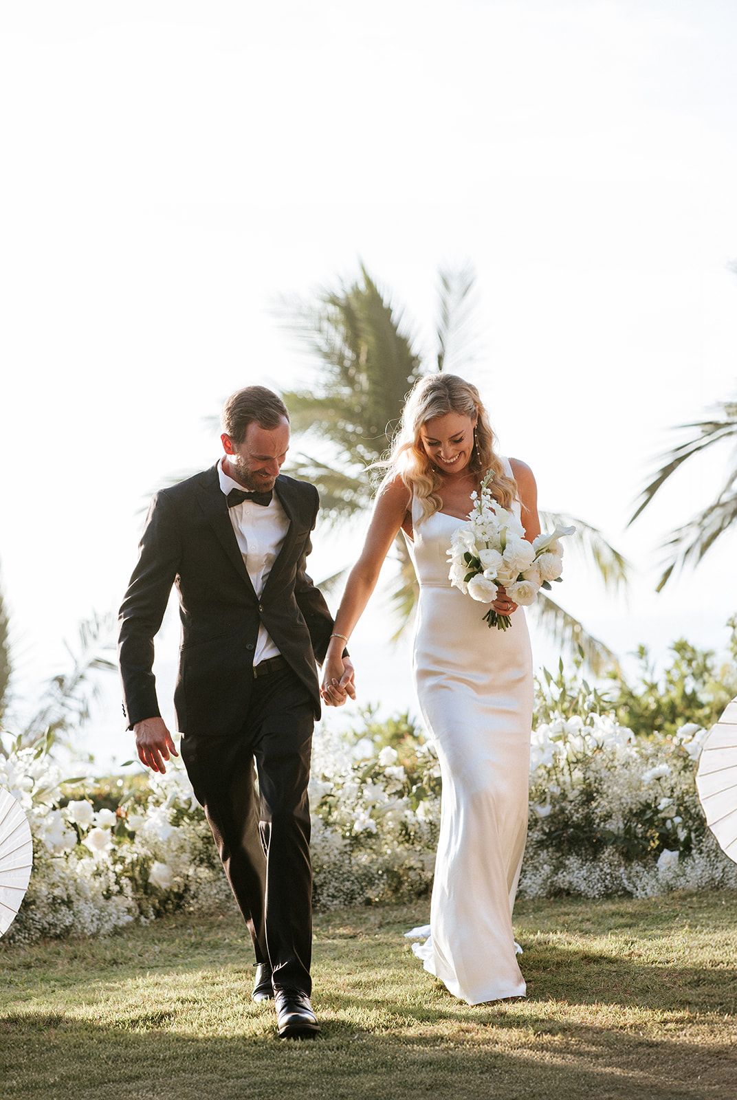 Bride and groom walking joyfully through a flower-lined garden after their ceremony in Puerto Vallarta, by José Carrillo Photography.