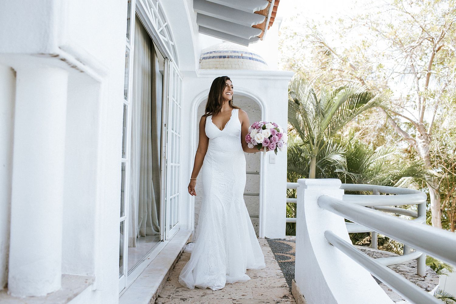 Elegant bride holding pastel roses steps onto a white villa balcony in Puerto Vallarta, captured by luxury photographer José Carrillo.