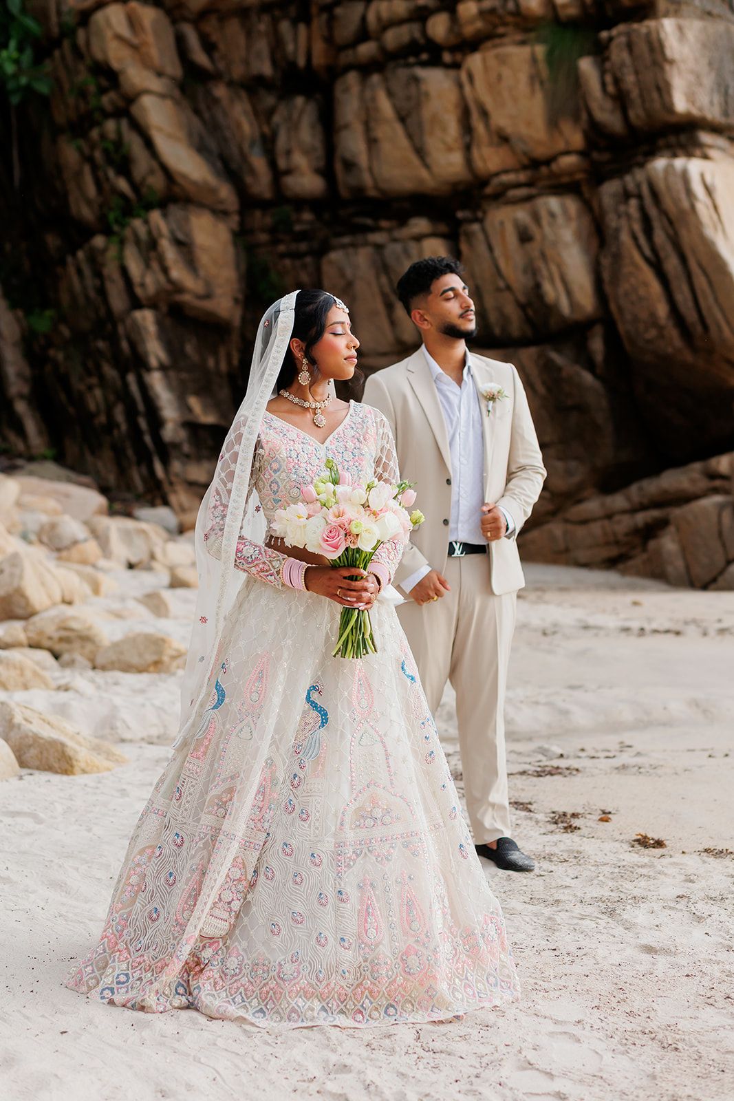 South Asian couple posing on the beach in Puerto Vallarta, bride in lehenga holding bouquet, by José Carrillo Photography.