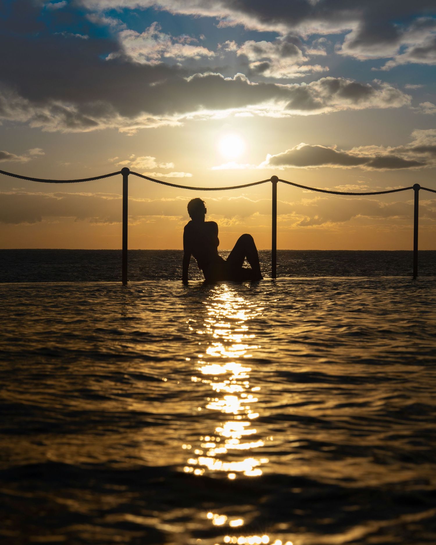 Silhouette of a person relaxing in the Bronte Rockpool at sunrise with golden reflections across the water and dramatic clouds overhead