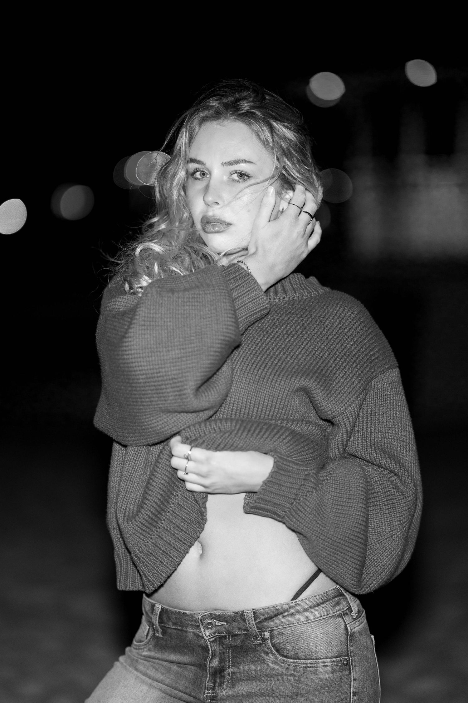 Black and white night portrait of a woman in a knitted sweater at Bondi Beach with blurred lights in the background.
