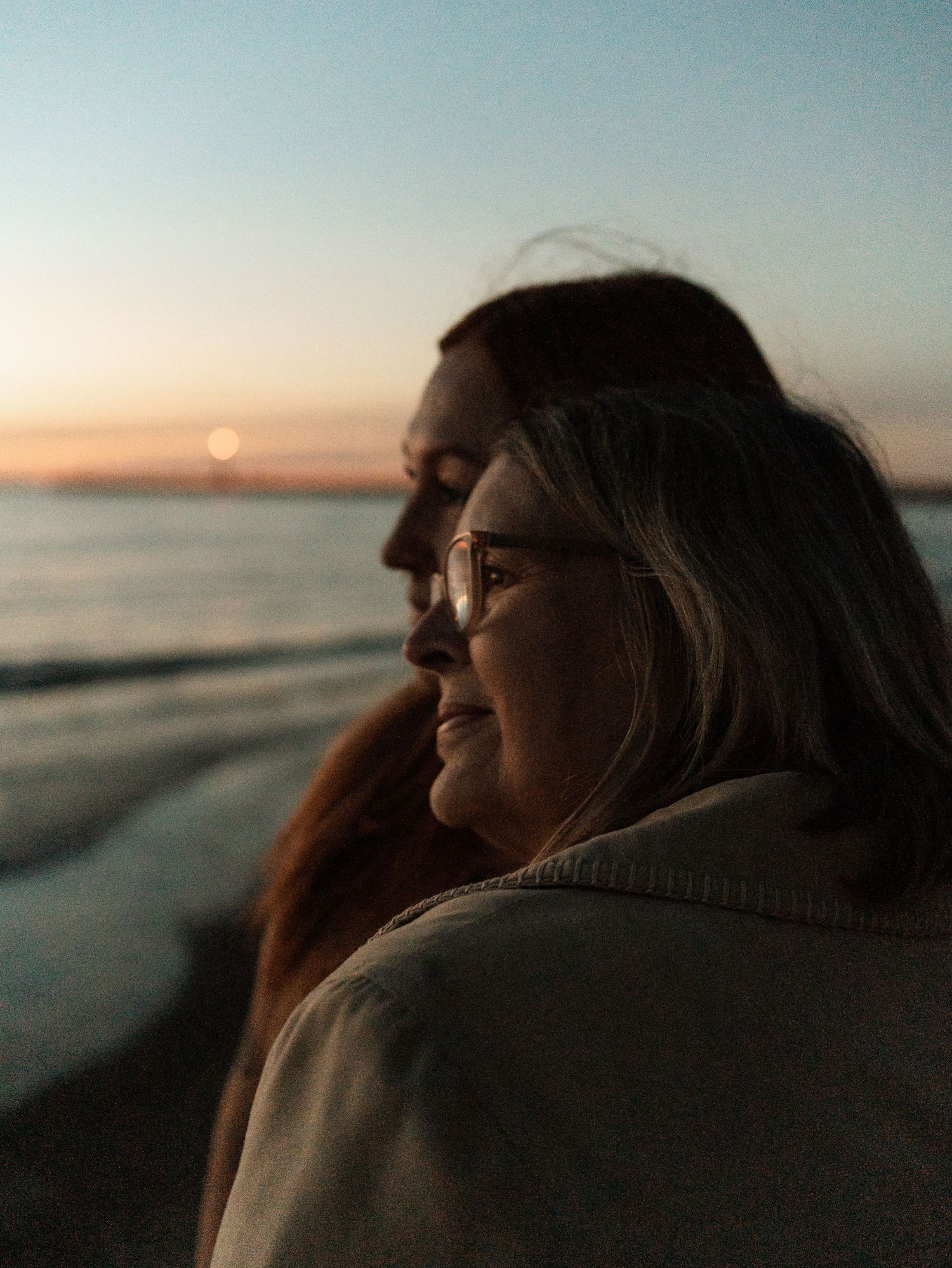 Two silhouettes sit together watching a peaceful sunset over calm ocean waters at dusk.