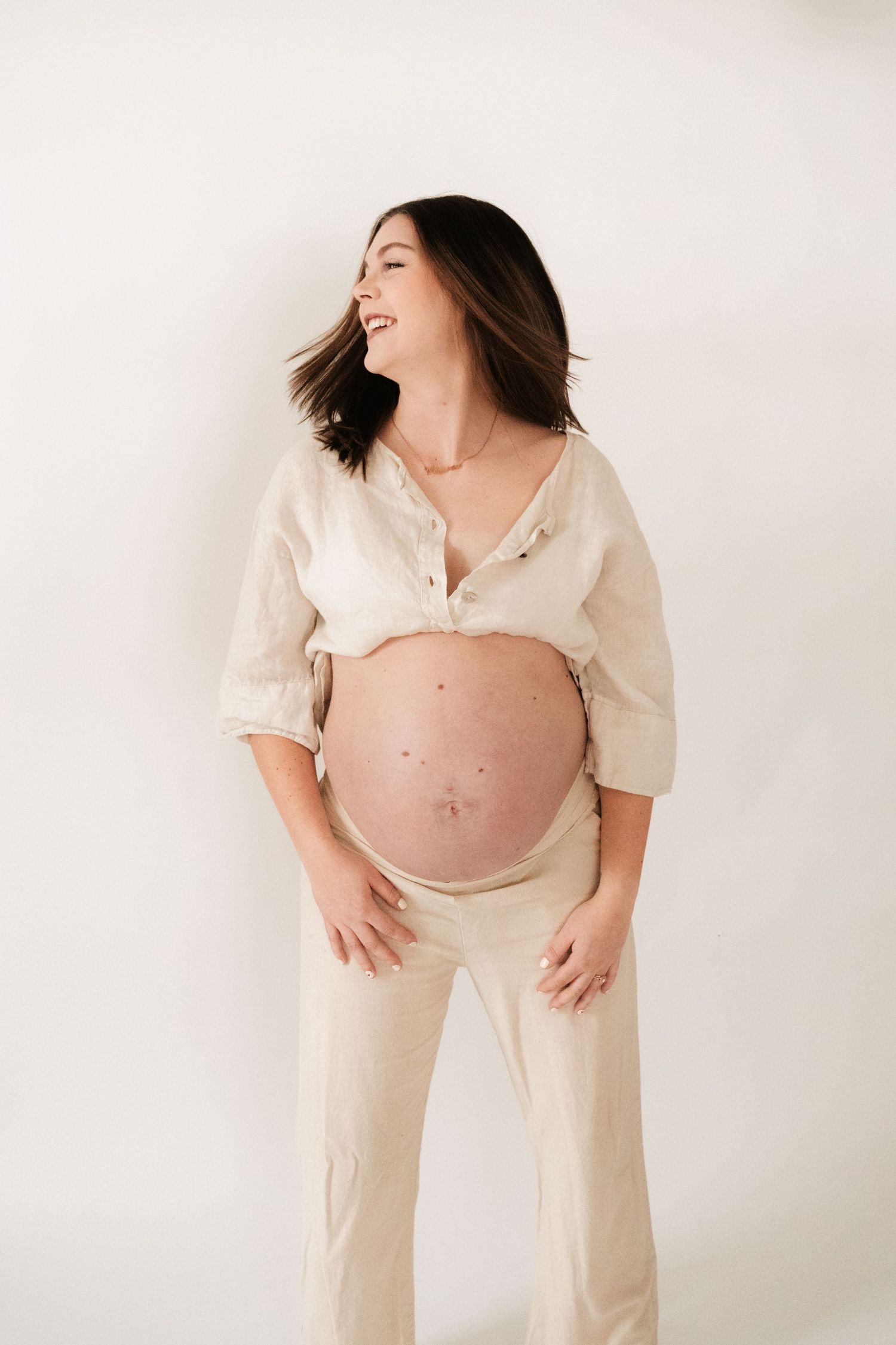 Expectant mother in cream-colored loungewear laughs joyfully during maternity photoshoot against white background.