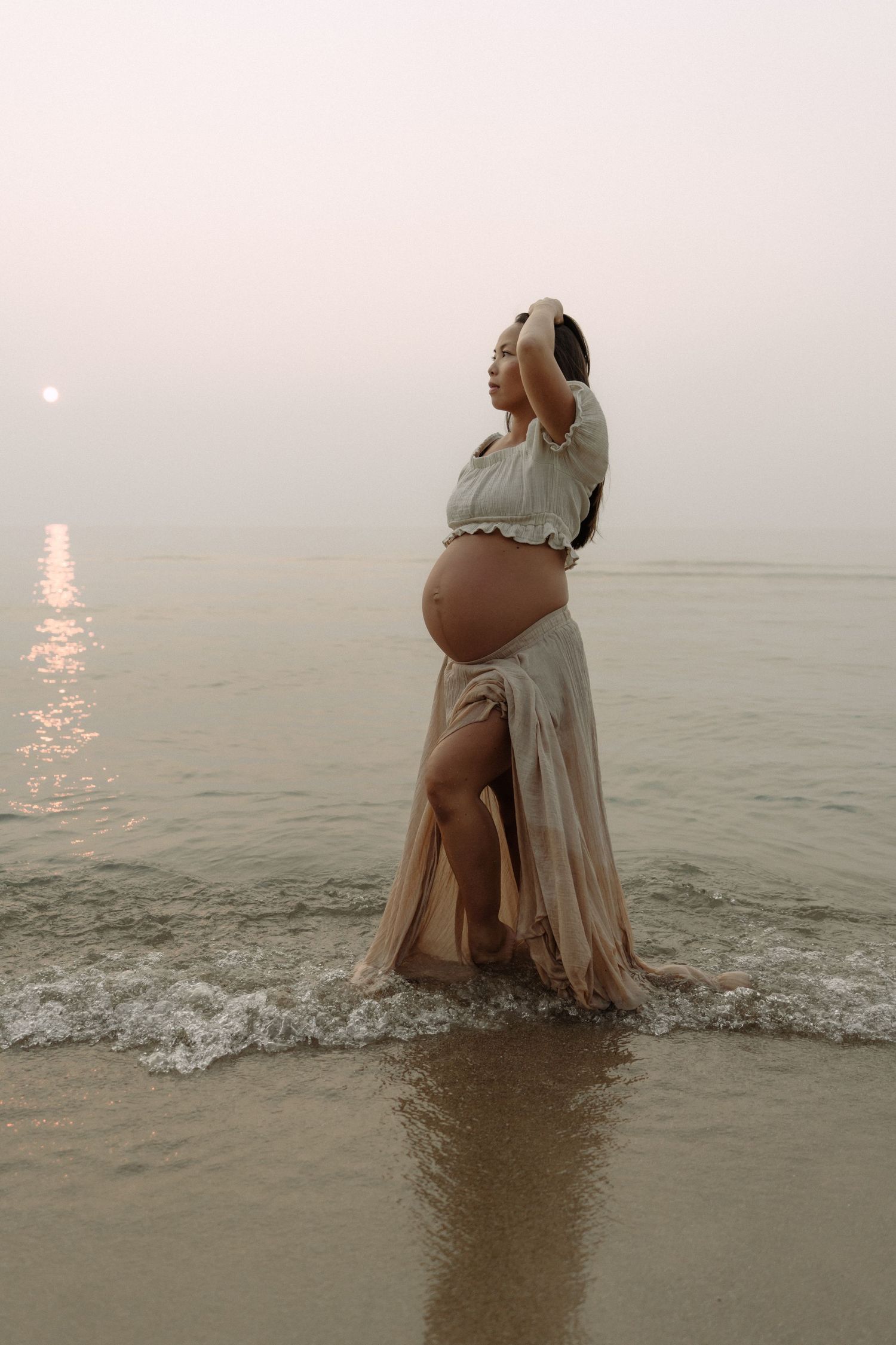 Expectant mother in flowing dress poses at sunset on misty beach with waves lapping at her feet.
