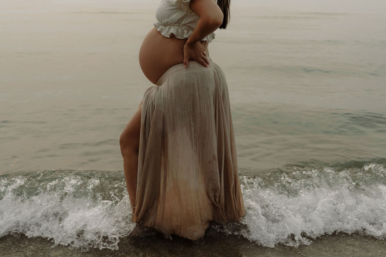 A pregnant figure in a flowing beige dress stands at the ocean's edge as waves crash around their feet.
