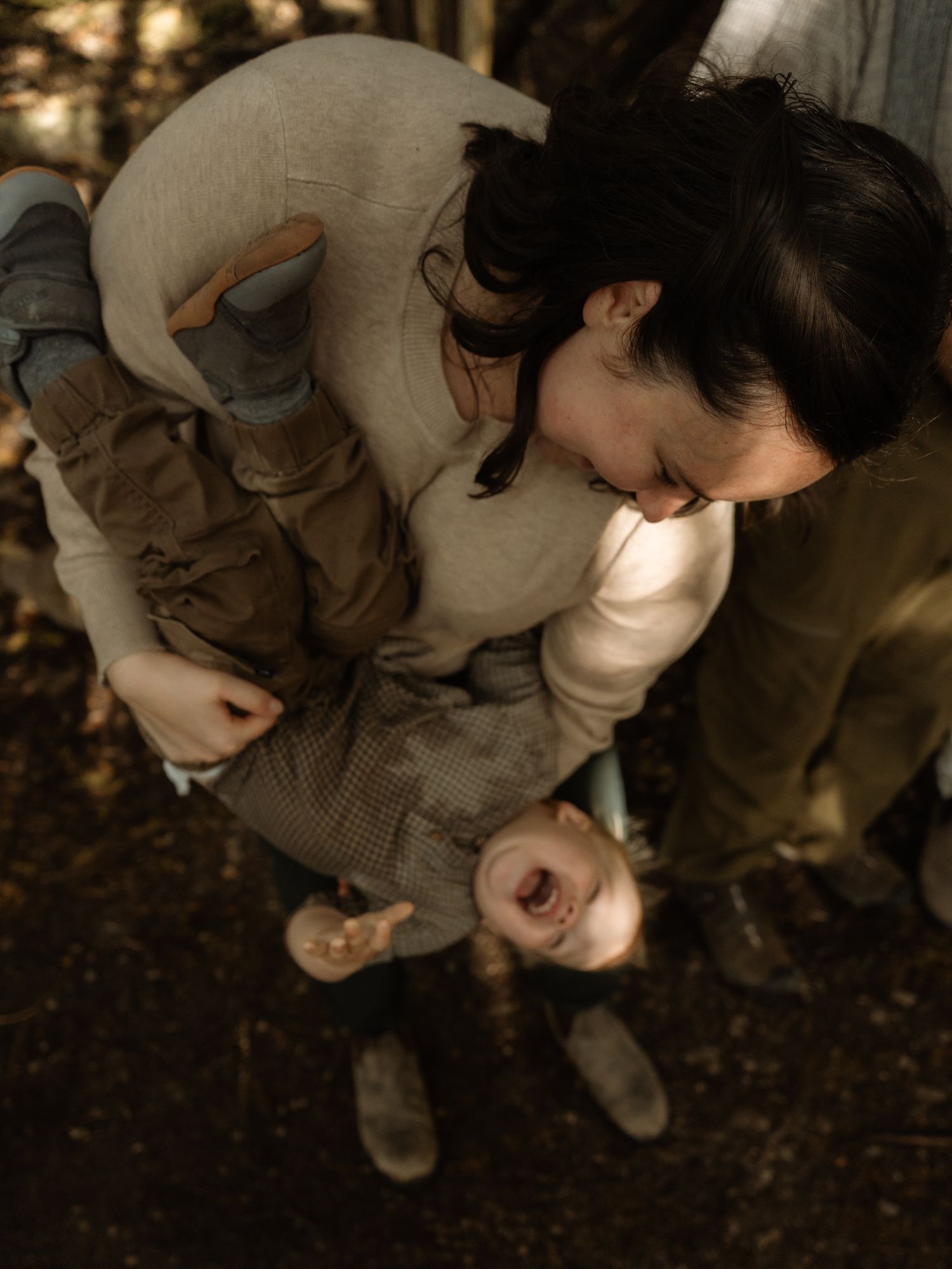 Mother and son in casual attire crouch down to examine something on the ground in a wooded outdoor setting.