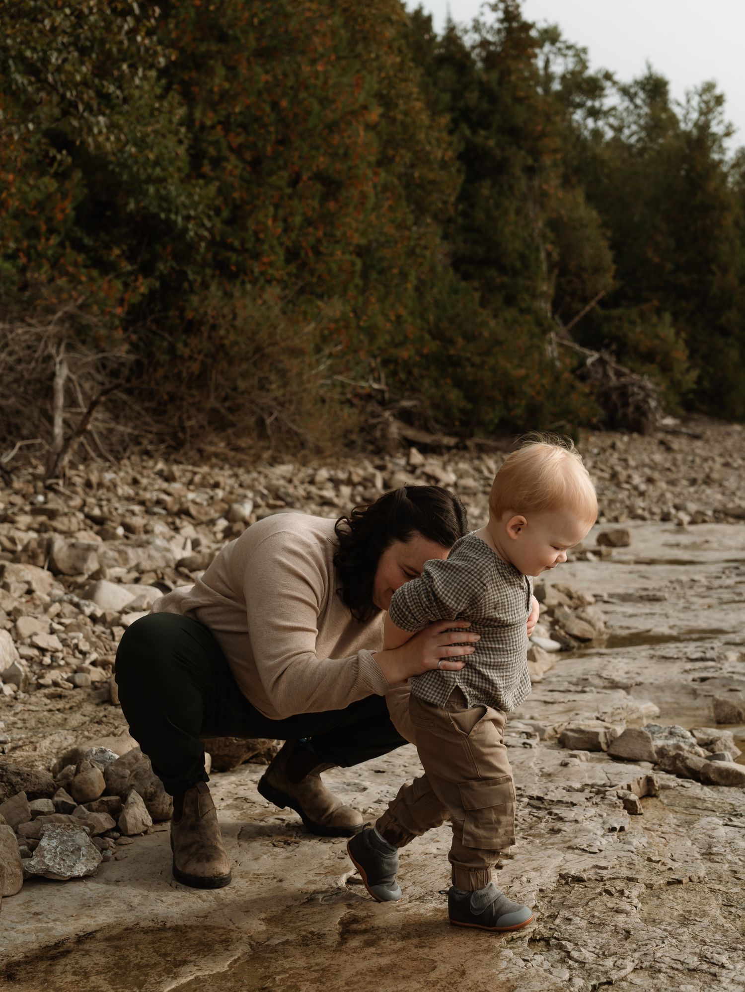 Two people explore a rocky riverbed together in matching tan outfits while searching for something on the ground.