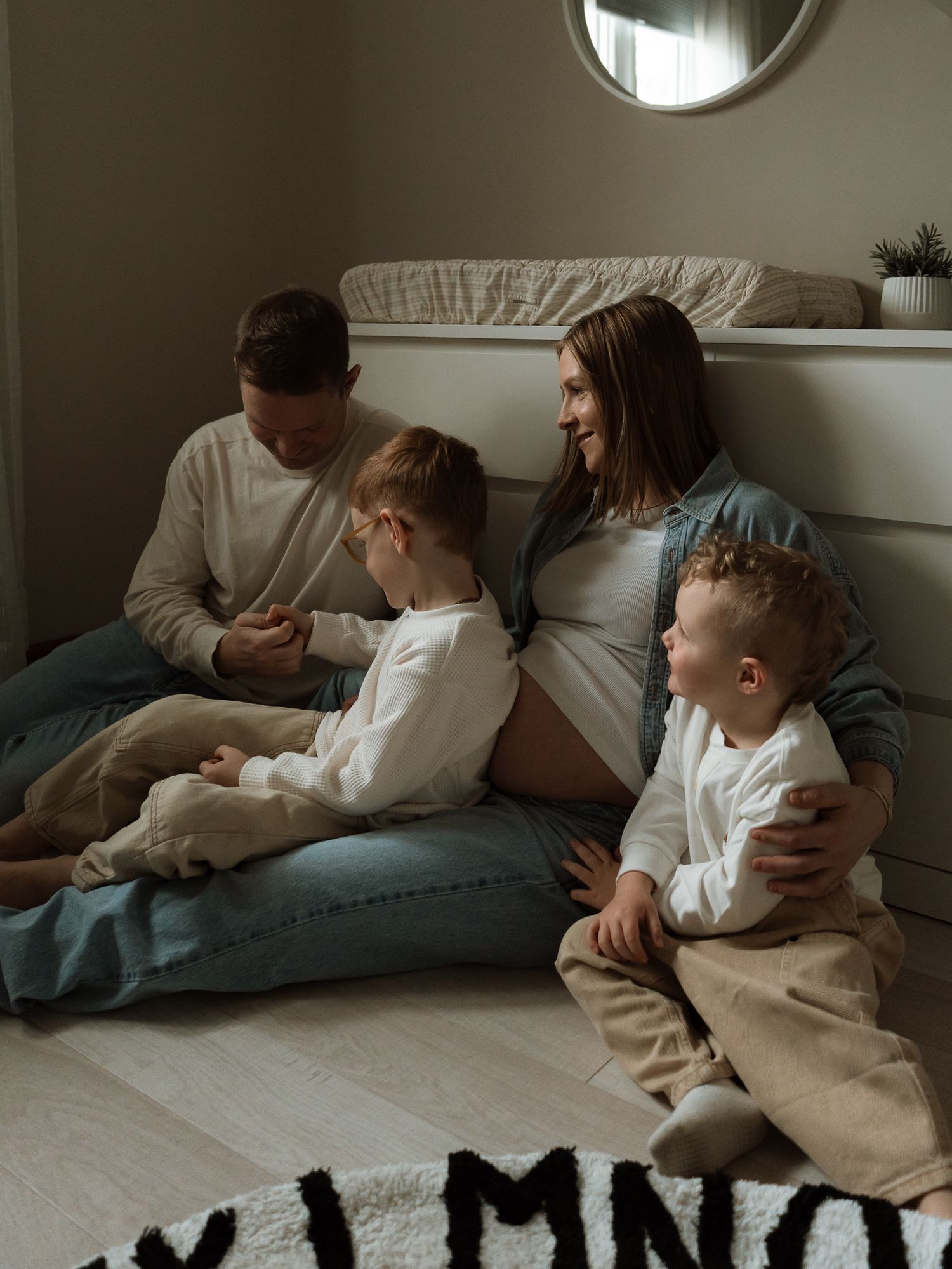 Pregnant woman sits on floor with husband and two young boys in cozy bedroom setting.