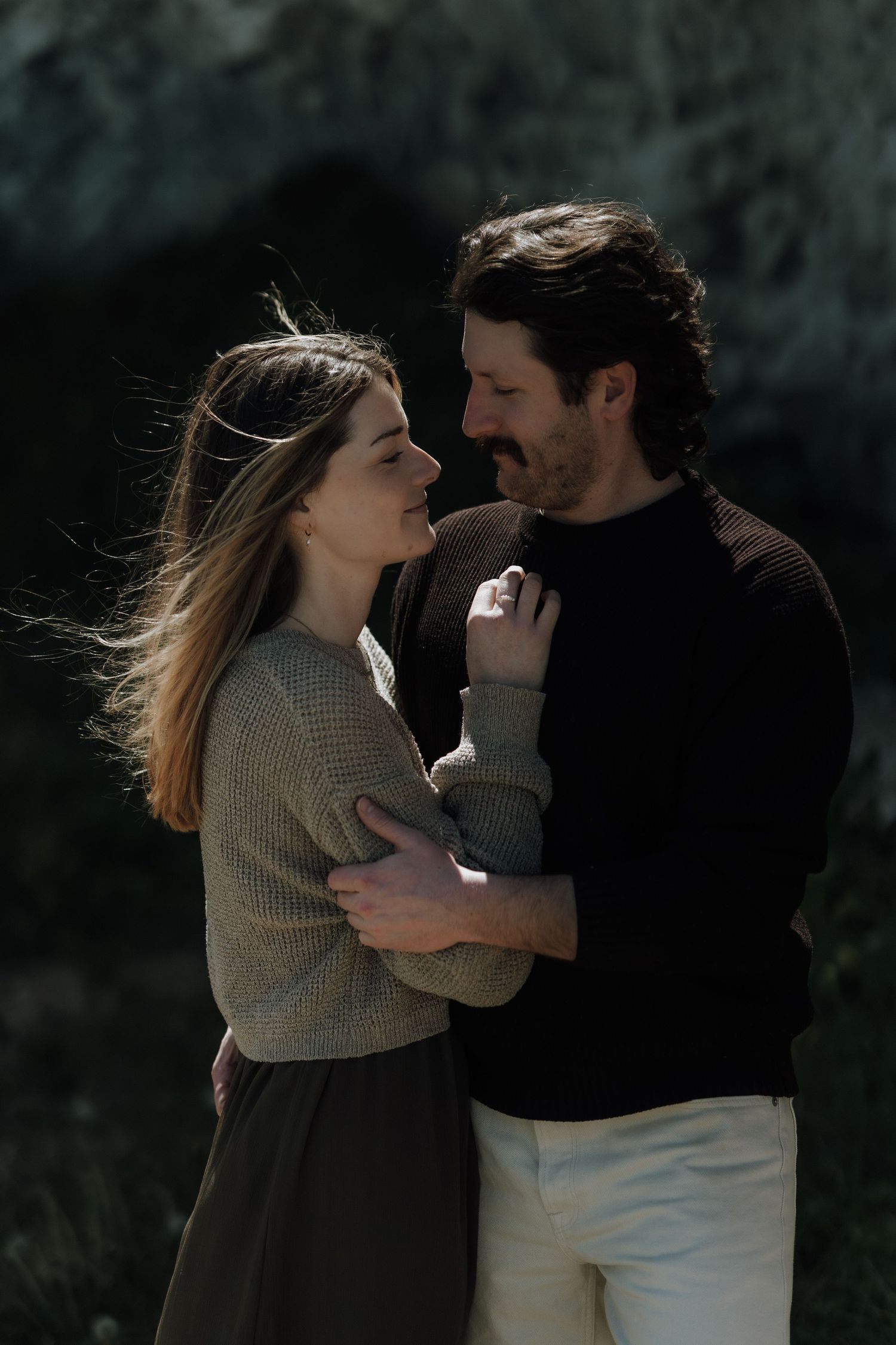 A couple embraces outdoors, the woman looking up lovingly at the bearded man in a black sweater, with wind blowing her hair.