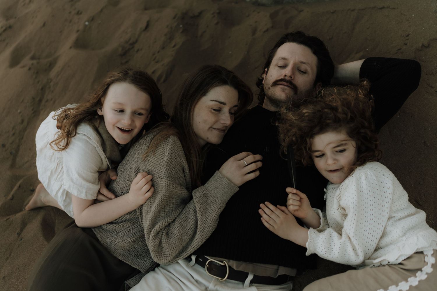Family of four lying together on sandy ground, laughing and embracing in a cozy outdoor moment.
