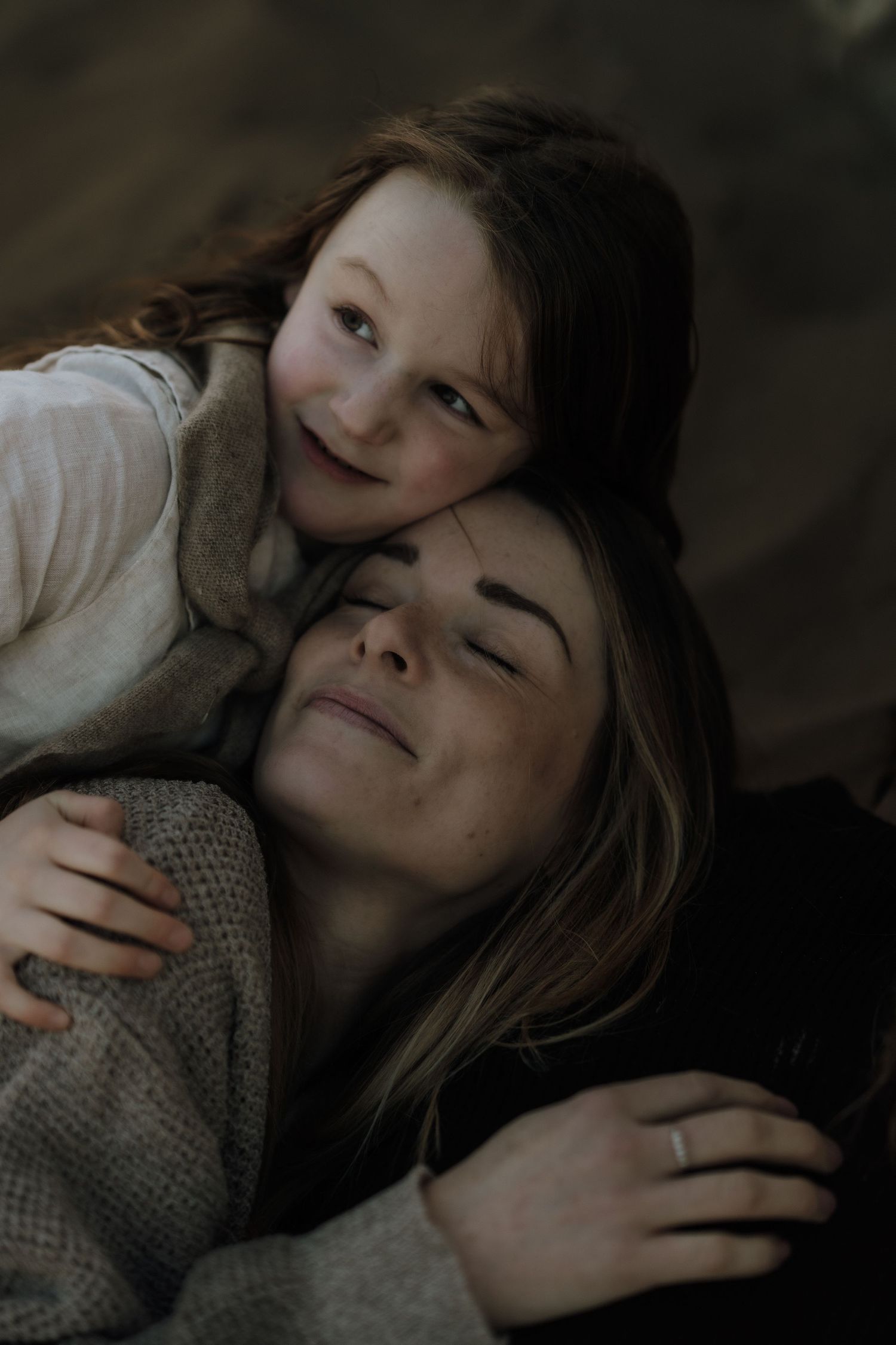 A young red-haired girl hugs a woman from behind, both sharing a tender moment in soft, muted tones.