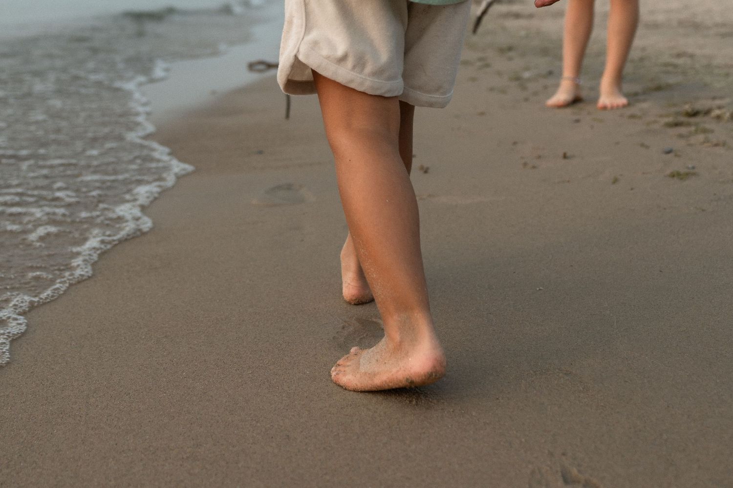 Bare feet walking along a sandy beach shoreline with gentle waves lapping at the shore.