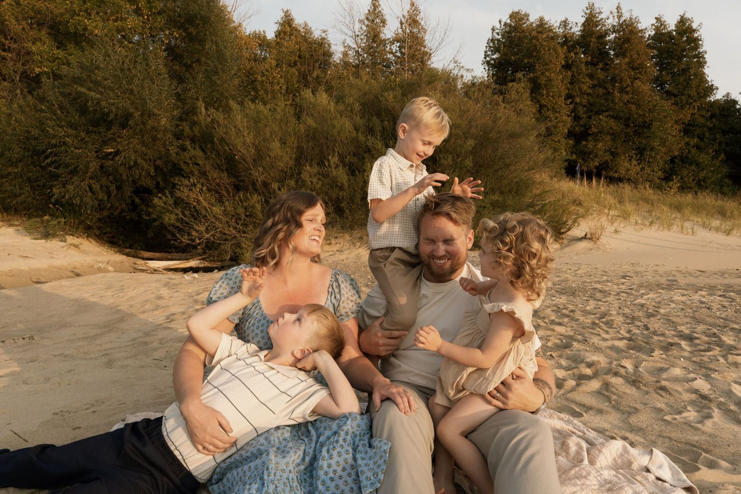 Family enjoying a relaxing evening together on a sandy beach at sunset with evergreen trees in the background.