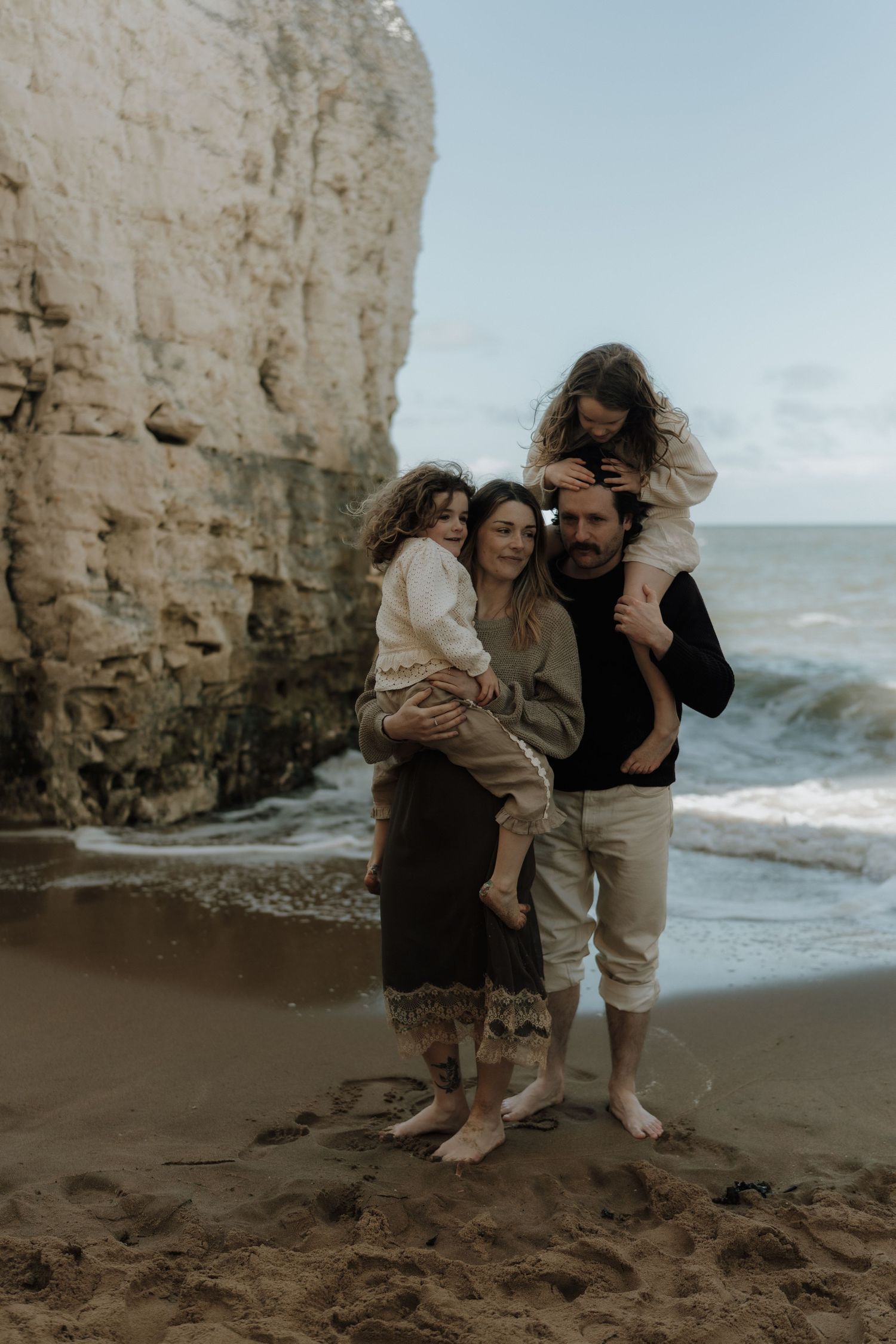 Family of four posing barefoot on sandy beach near rocky cliffs, parents each holding a child, ocean waves in background.