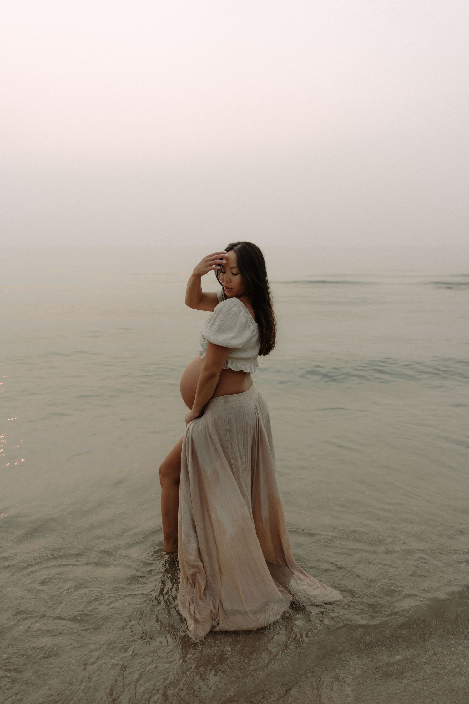 A woman in a flowing white dress stands in shallow ocean water at sunset creating a dreamy coastal scene.