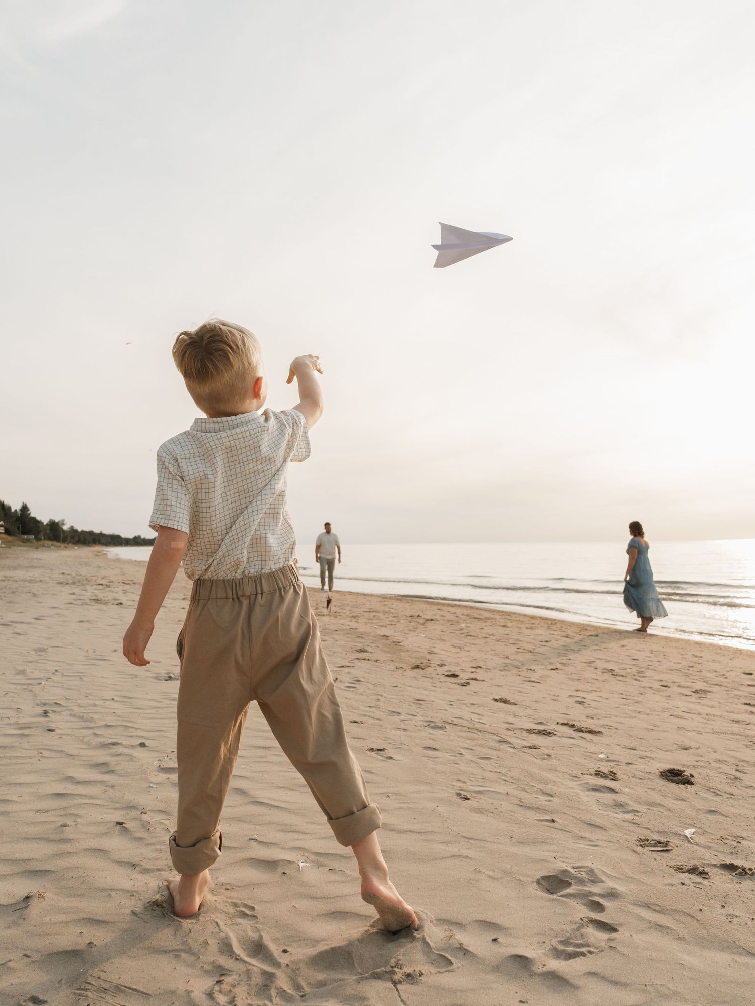 Child throwing paper airplane on a peaceful beach at sunset while people walk along the shoreline.