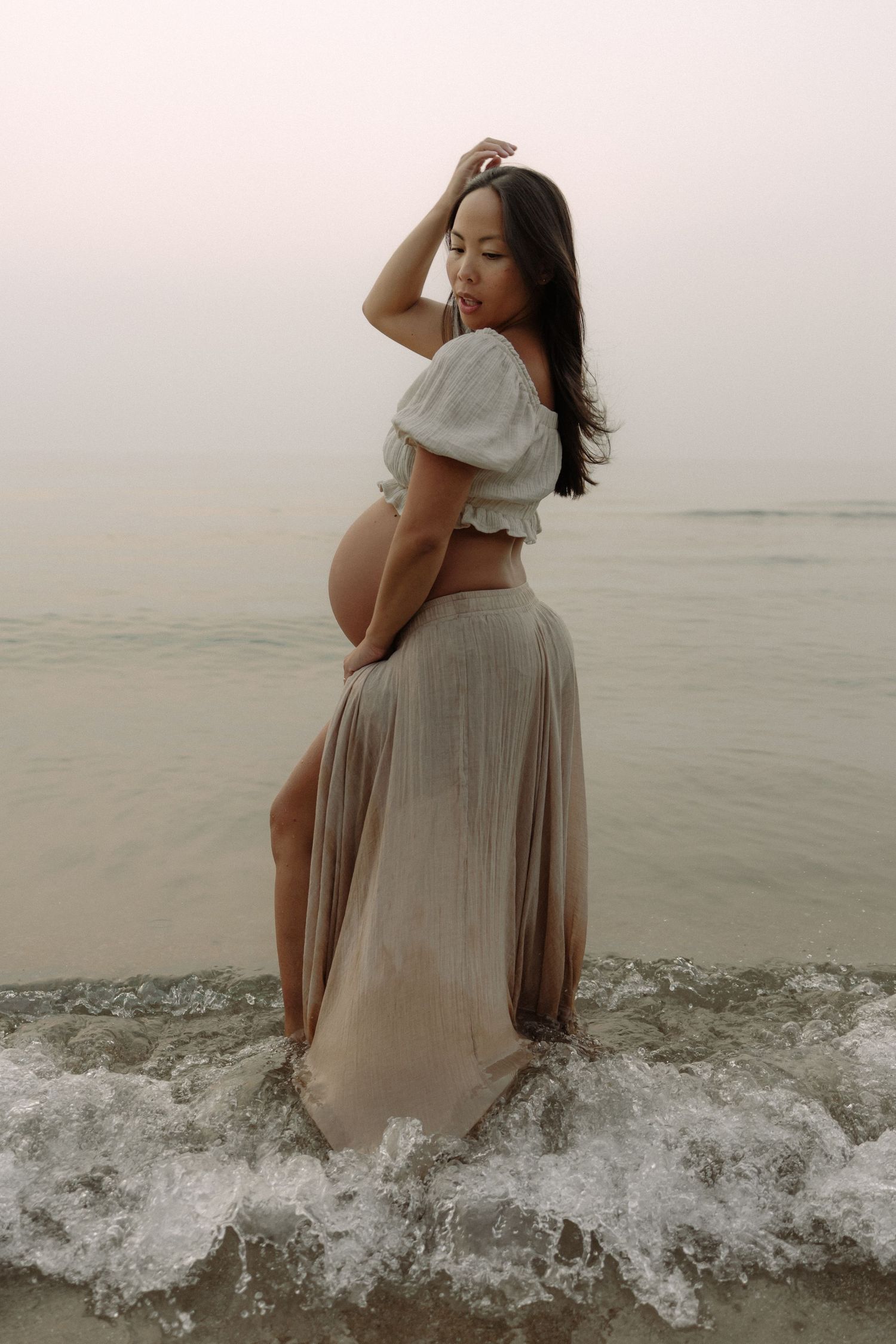 A pregnant woman in a flowing skirt and crop top poses at the beach during sunset as waves crash around her feet.