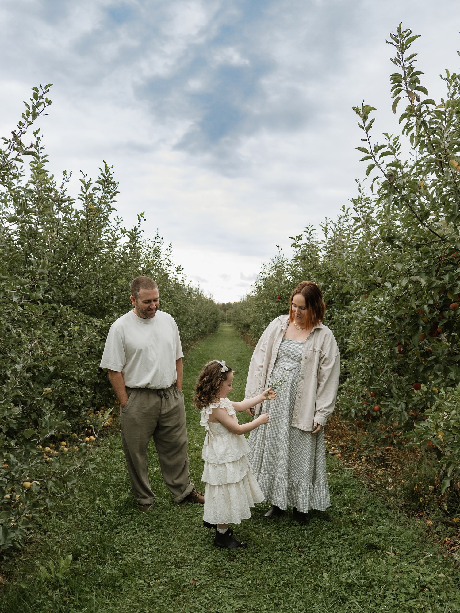 A family stands together in a lush apple orchard on a cloudy day wearing white and neutral colored clothing.