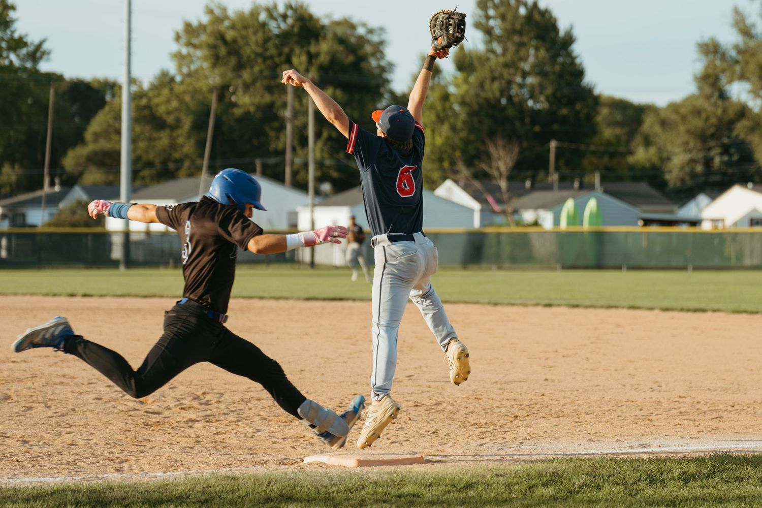 Baseball player #6 catches ball at first base while runner slides in during youth league game on sunny day.