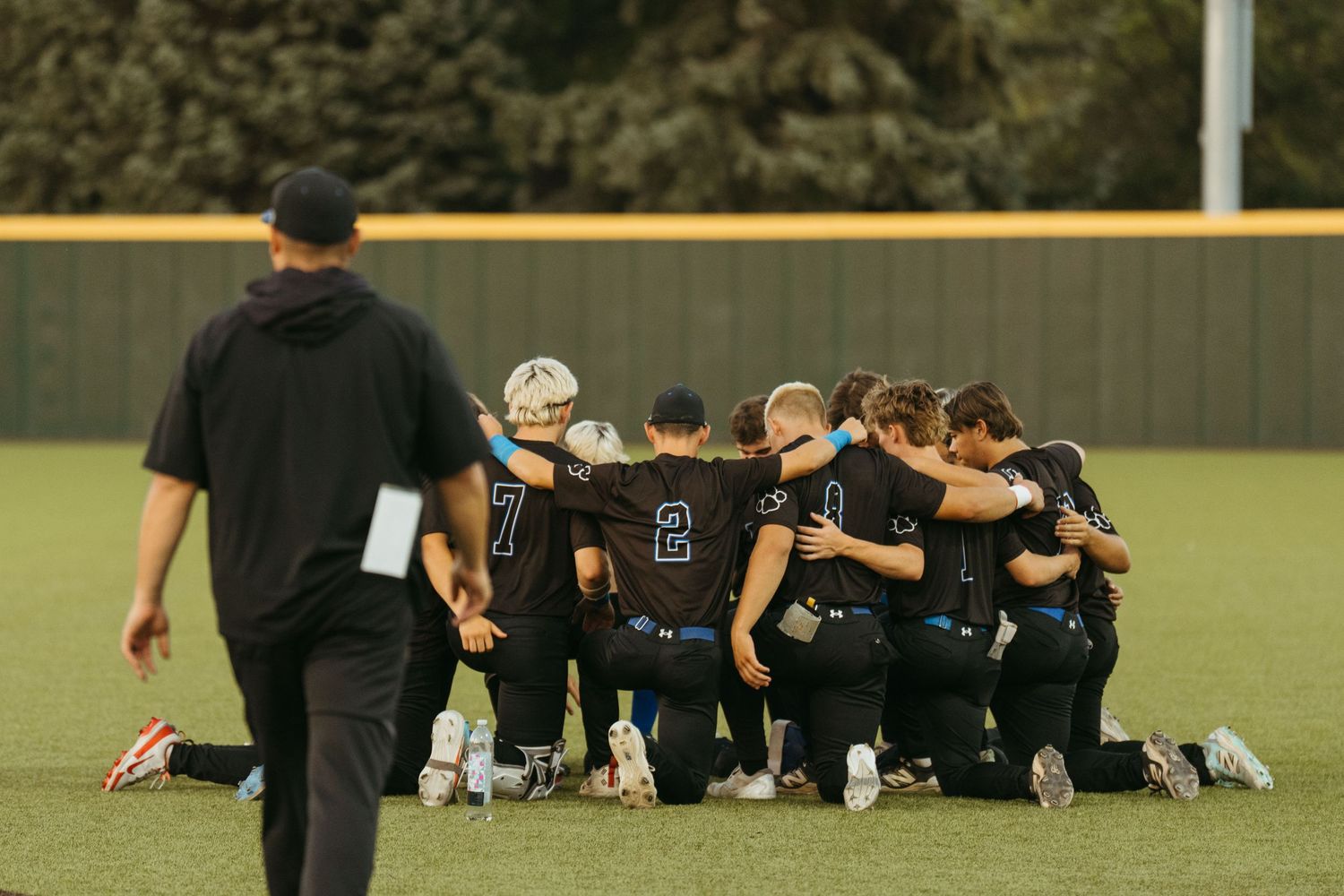 Baseball team huddles together on field in dark uniforms during a game, with coach walking nearby.