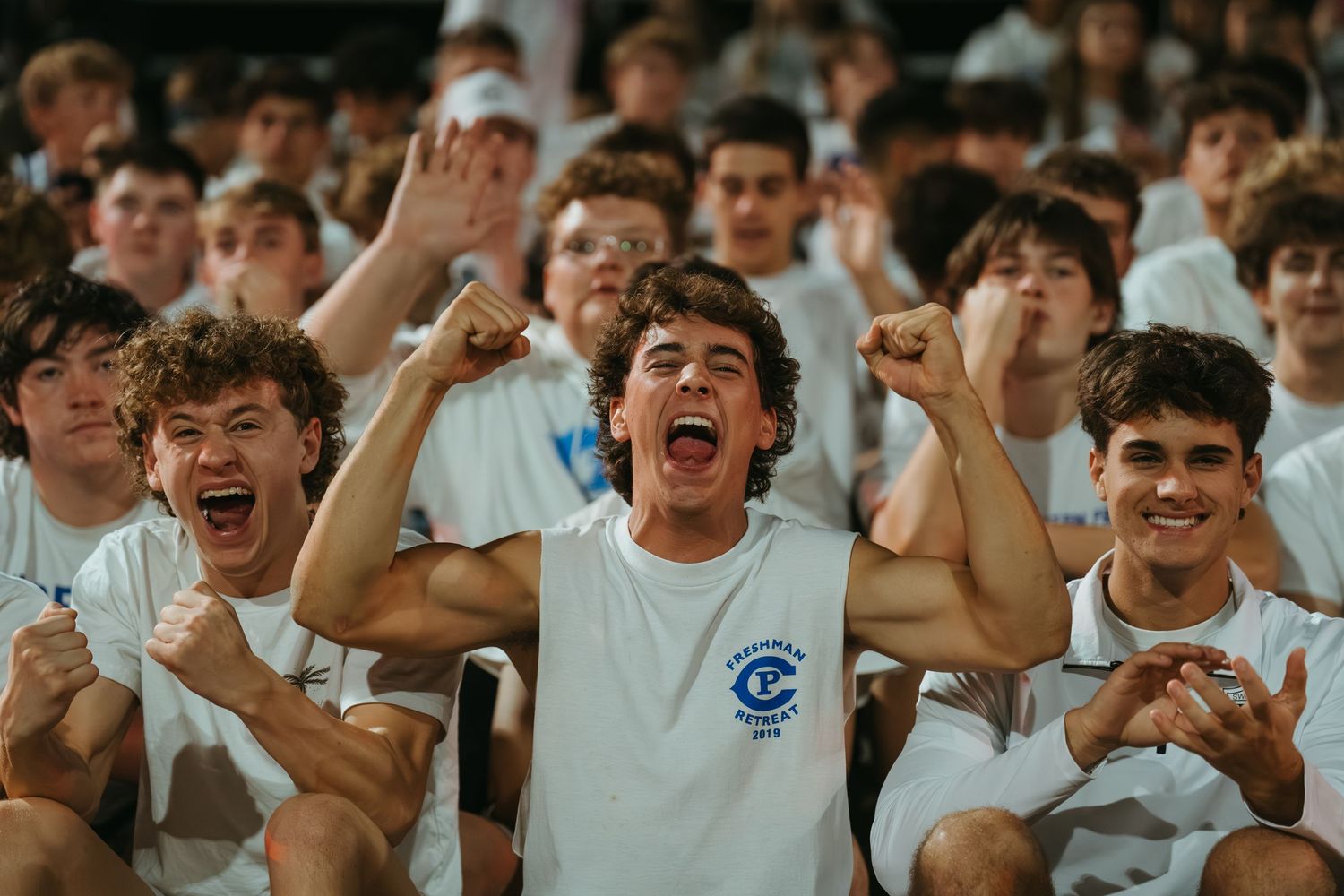 Excited young men in white t-shirts cheering enthusiastically at a school pep rally or sporting event.