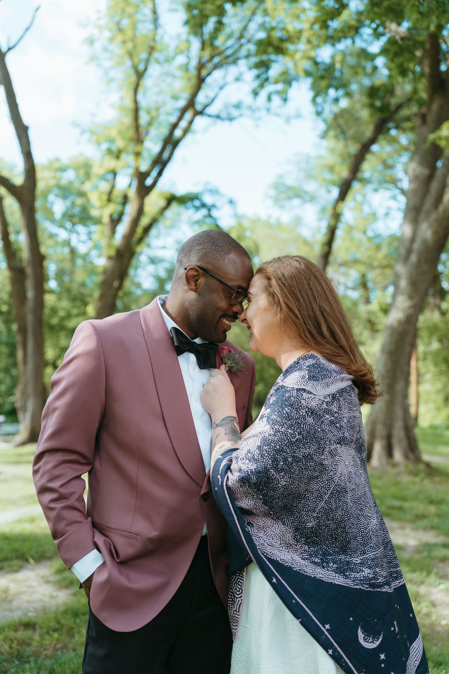 Interracial couple in romantic pose outdoors, man in pink tuxedo jacket, woman in celestial wrap, surrounded by trees.