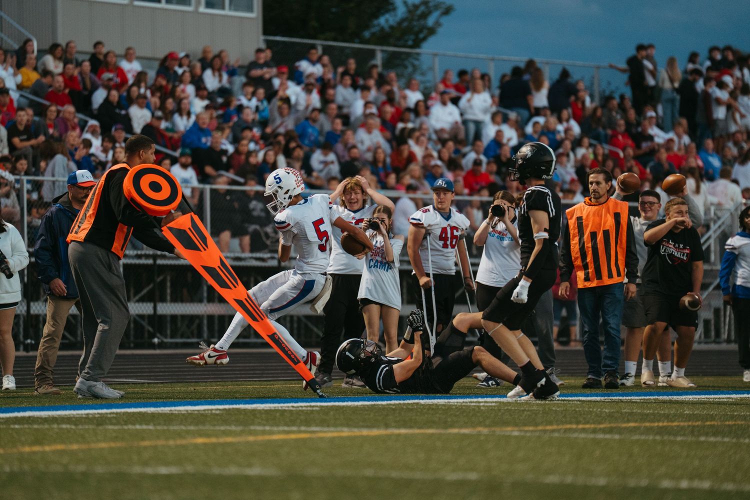 High school football players in red and white uniforms run during a game with referees and packed bleachers in background.
