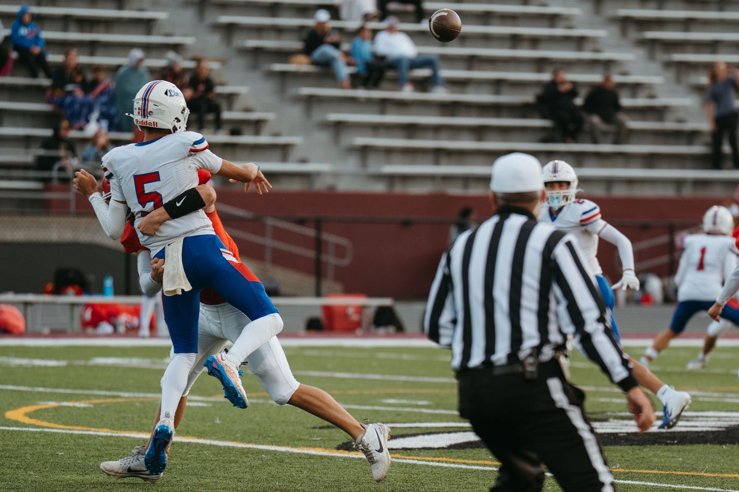 Football player #5 throws a pass during a competitive high school or college football game on a stadium field.