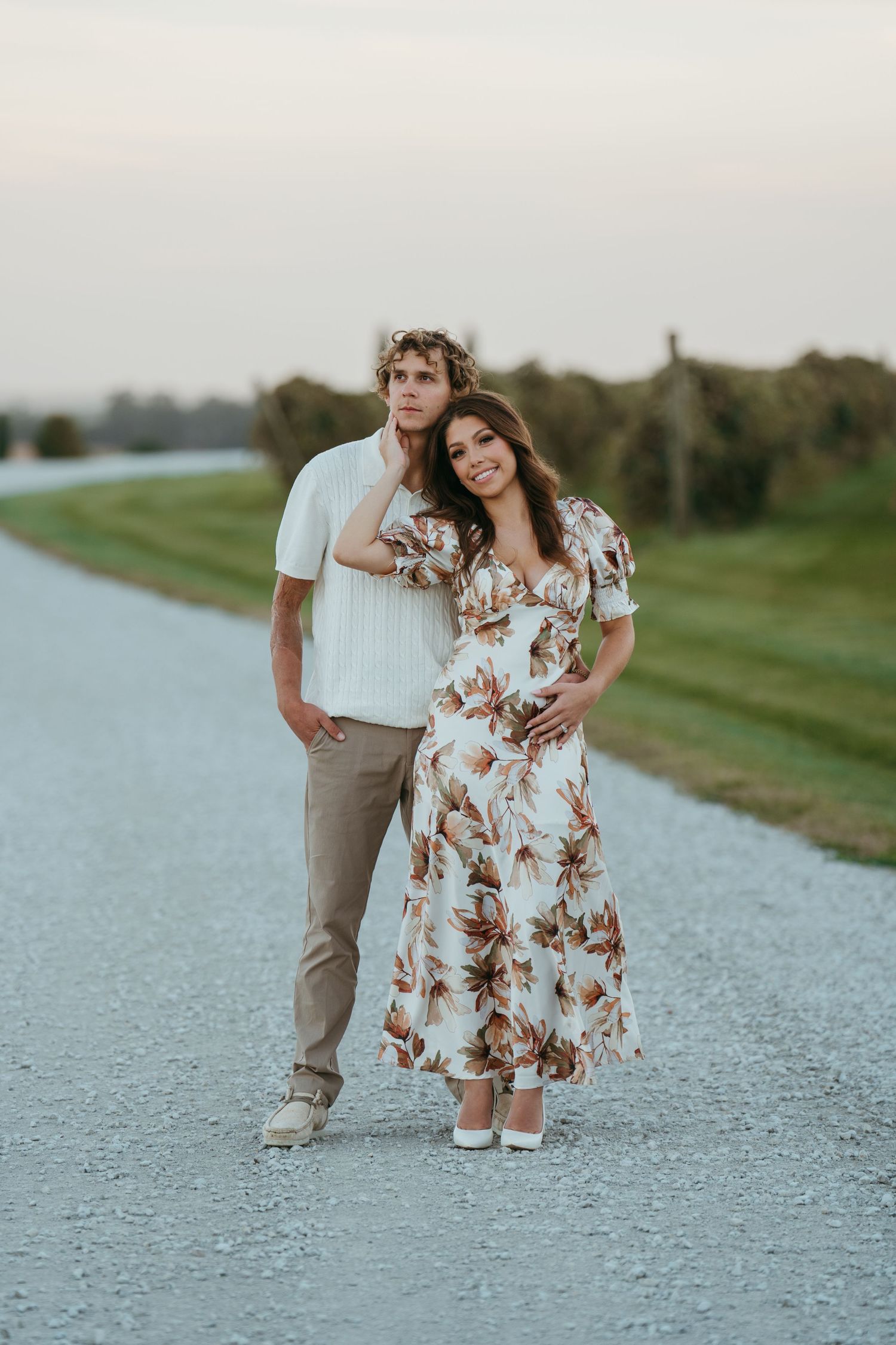 A couple stands together on a gravel path at sunset, wearing casual summer attire including a floral dress and white shirt.