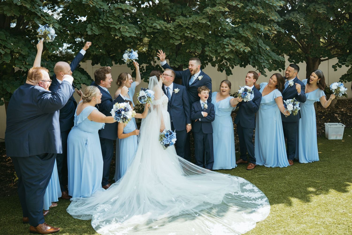 Wedding party in blue dresses and navy suits celebrate with bride and groom on sunny outdoor lawn.