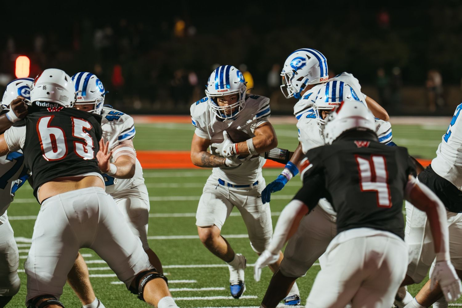 Football players in blue uniforms run through defenders in a dark, action-packed night game under stadium lights.