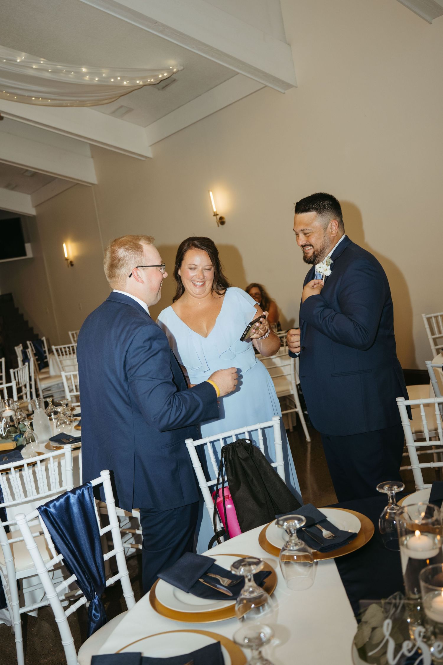 Three wedding guests in formal attire laughing together at a elegantly decorated reception venue with white chairs.