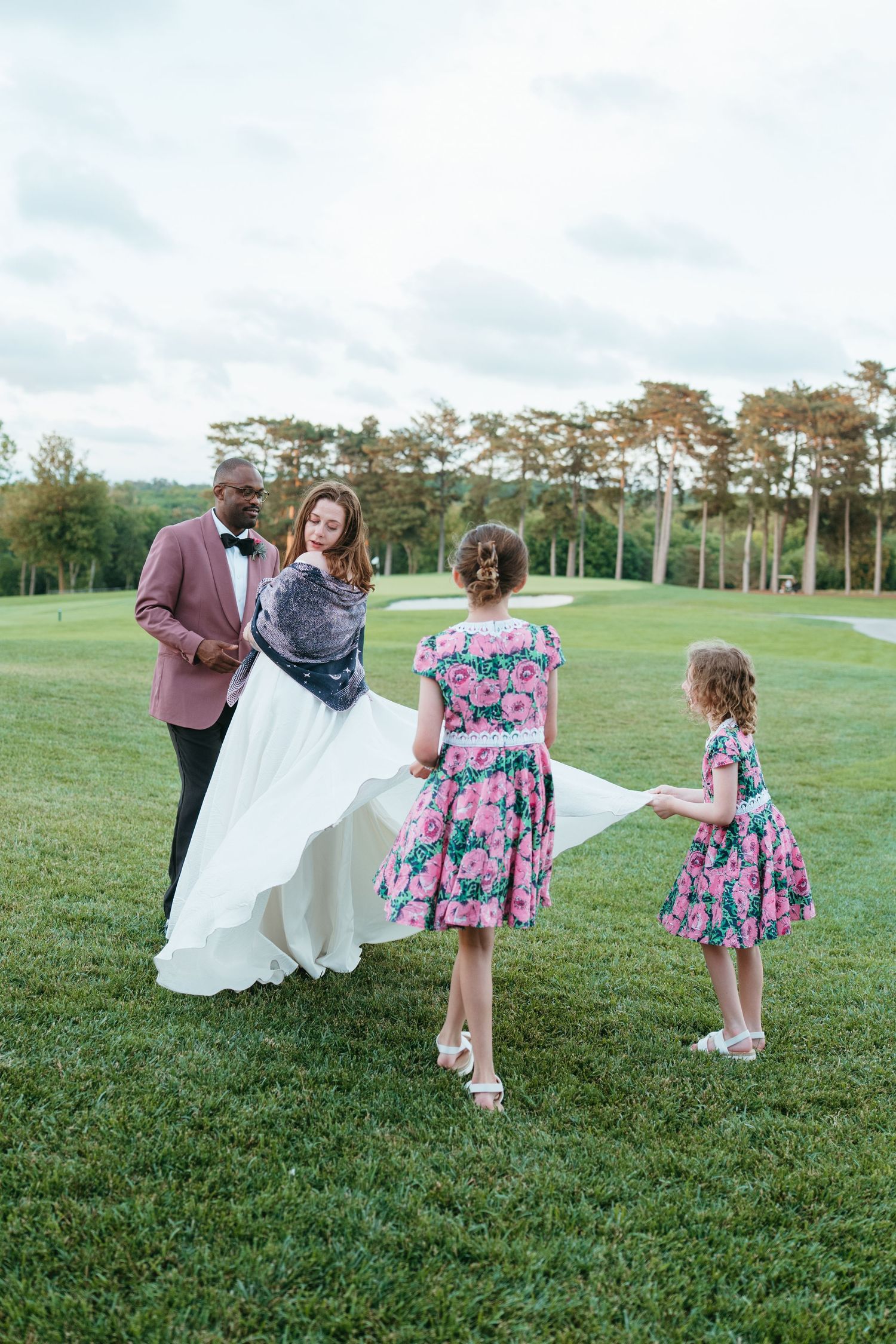 Interracial wedding couple with two girls in floral dresses on a green golf course lawn.