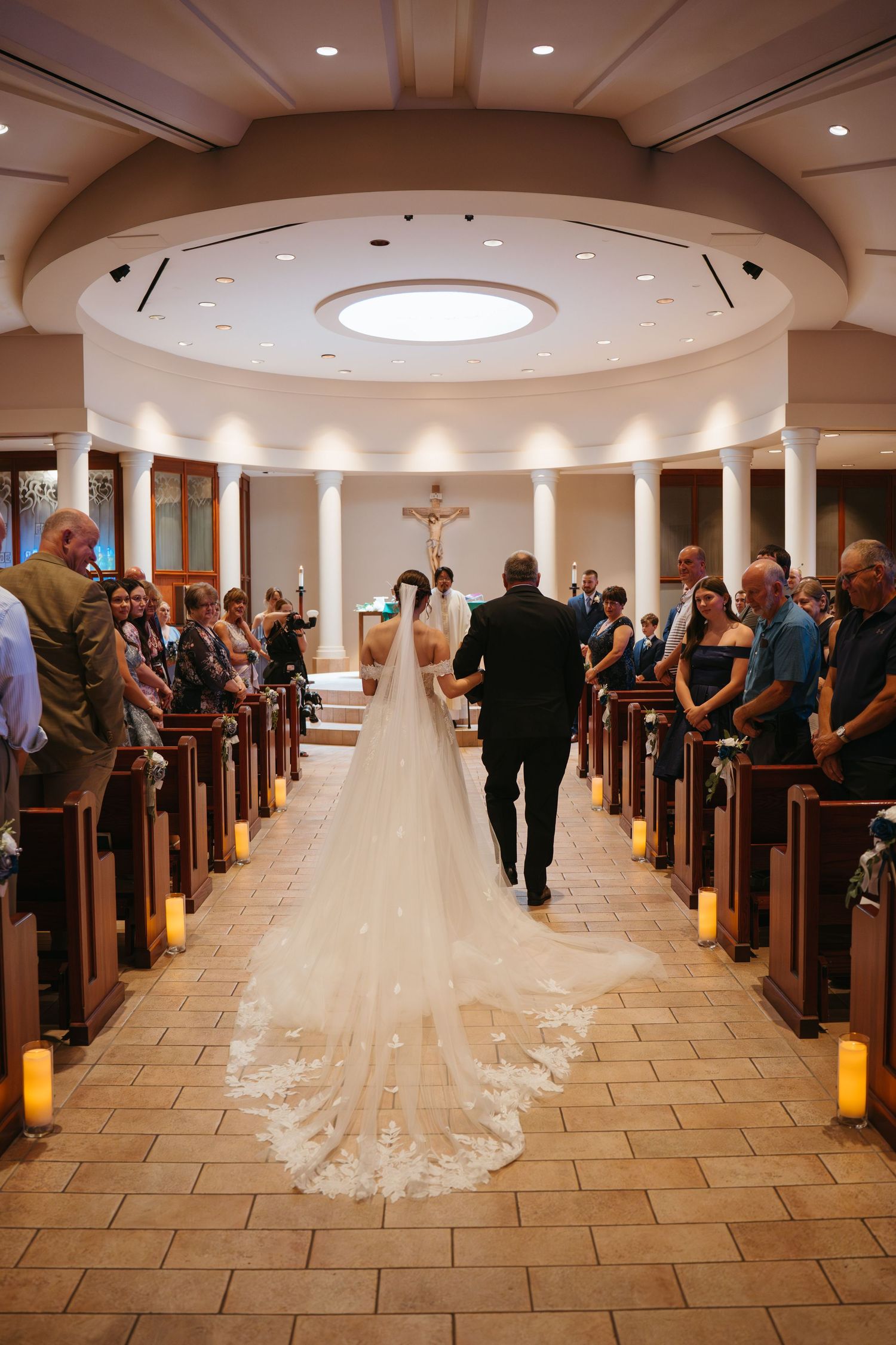 Bride walking down the aisle with escort in elegant Catholic church, long lace veil trailing, guests seated in wooden pews.