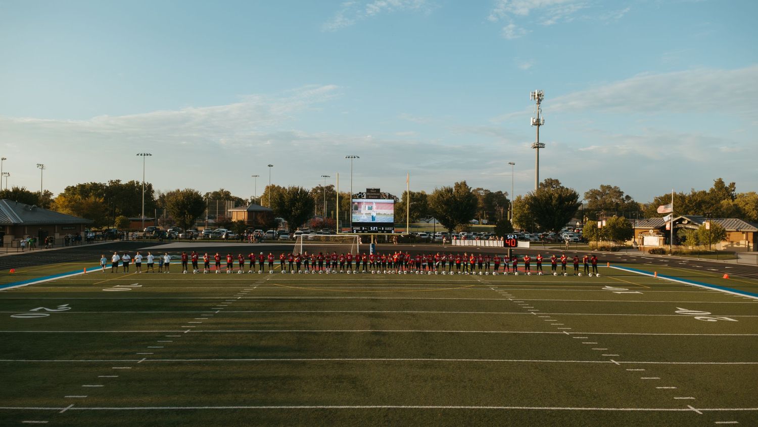 Marching band lined up on a high school football field at dusk with scoreboard and trees in background.