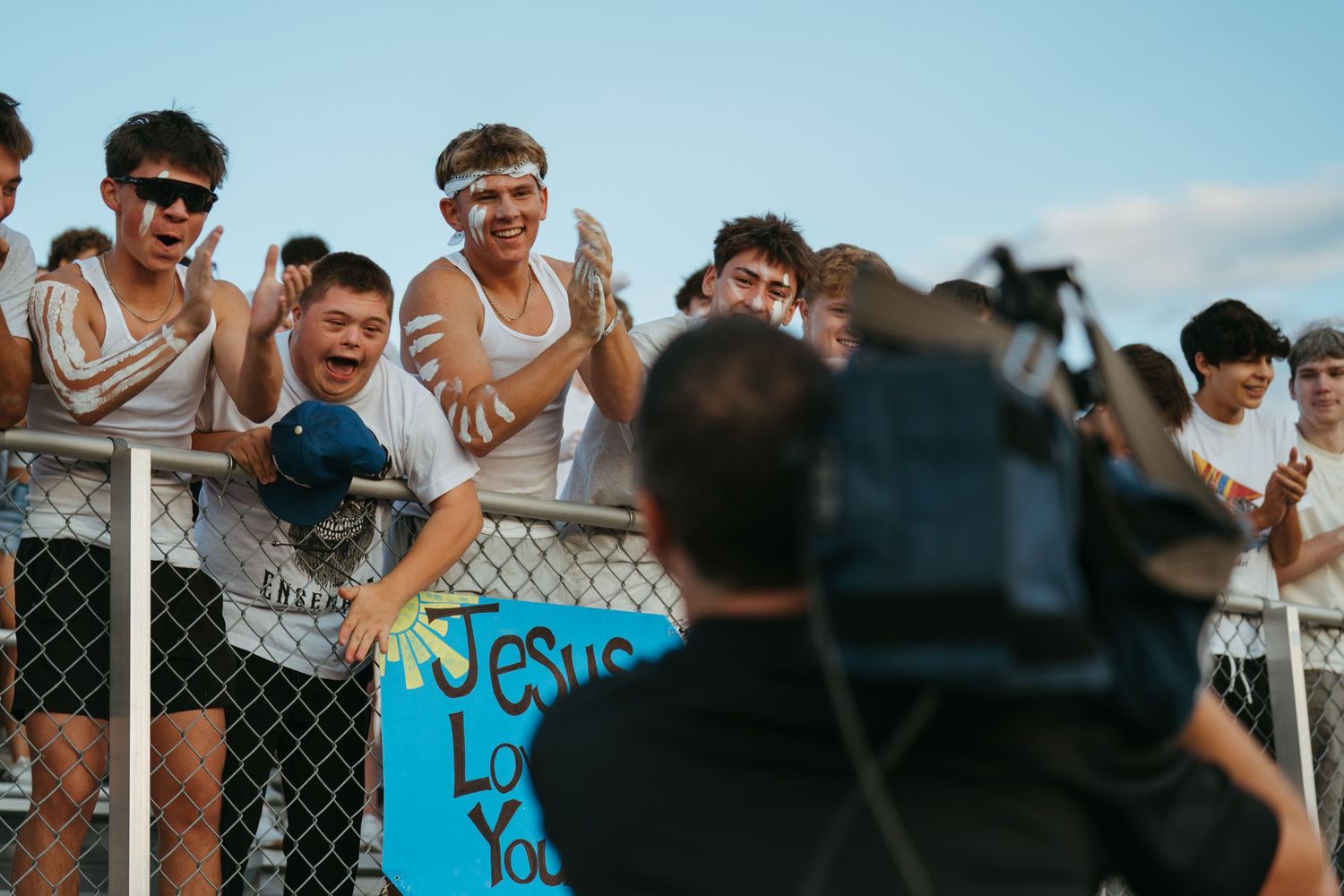 Excited teen fans cheer at a sports event holding a Jesus Loves You sign while being filmed by a cameraman.