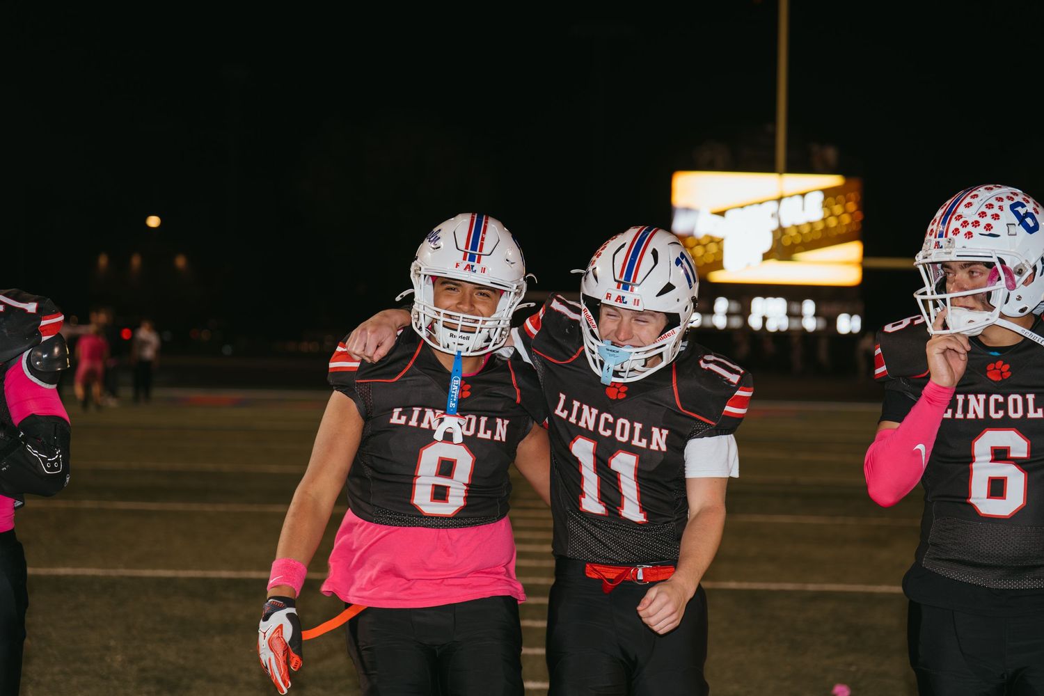 Lincoln football players #8 and #11 in pink jerseys celebrate on the field during a night game.