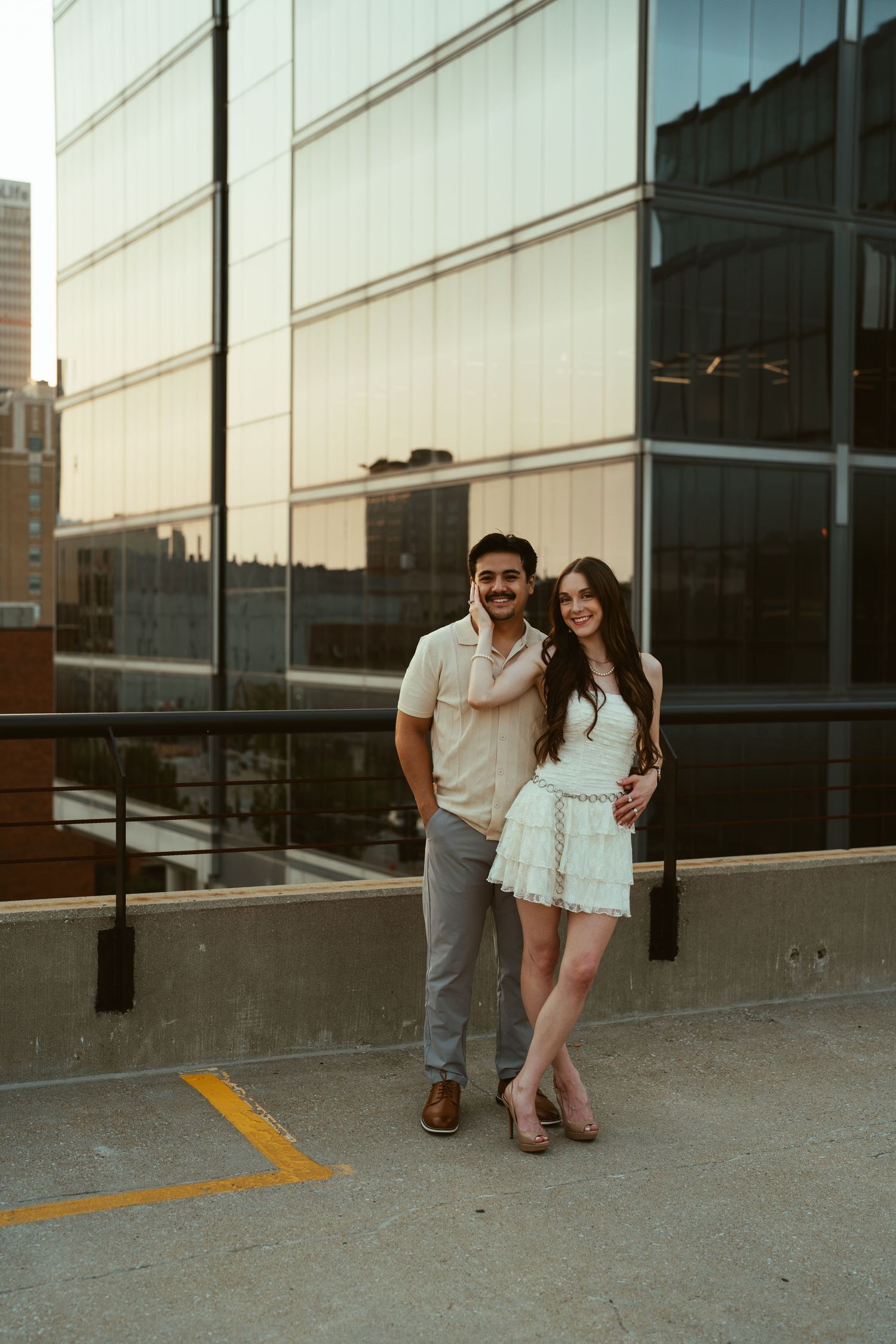 A couple poses together against a modern glass building exterior at sunset.