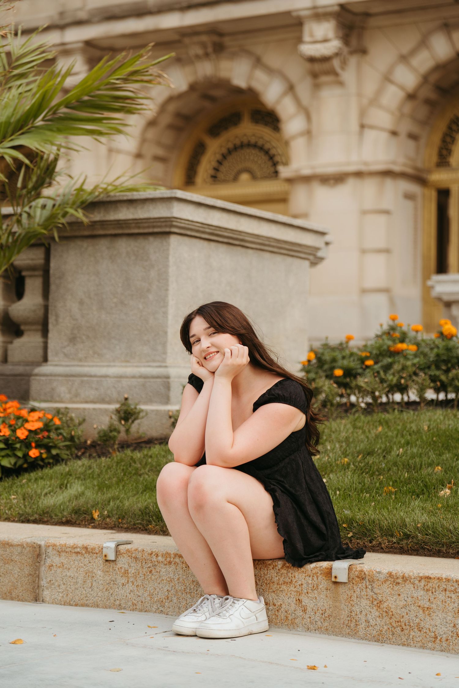 Person in black dress and white sneakers sits curled up on stone steps near orange flowers and palm trees.