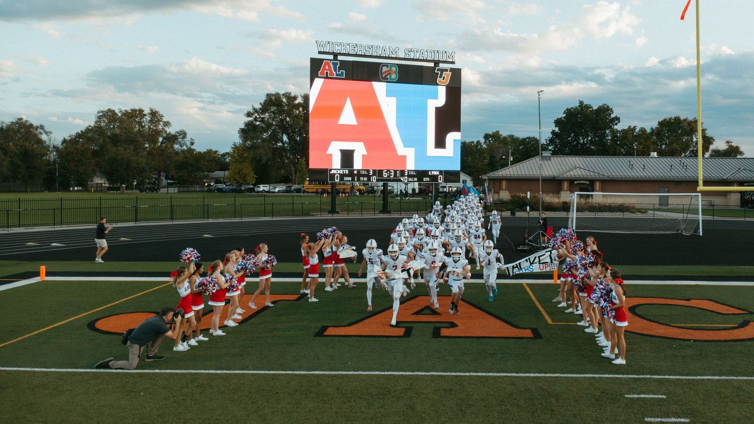 Football team runs onto field through cheerleader tunnel under large LED scoreboard at night game.