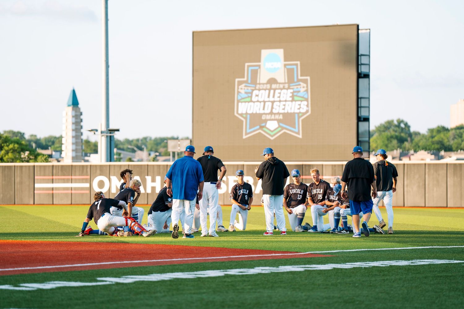 Baseball team gathers on field at College World Series venue under large scoreboard on sunny day.