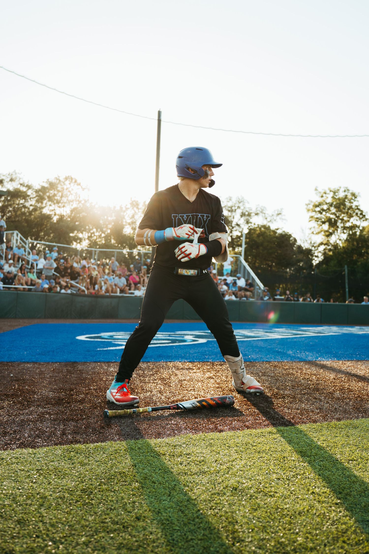 Female softball player in blue helmet stands ready at bat during a sunny evening game with crowd in background.