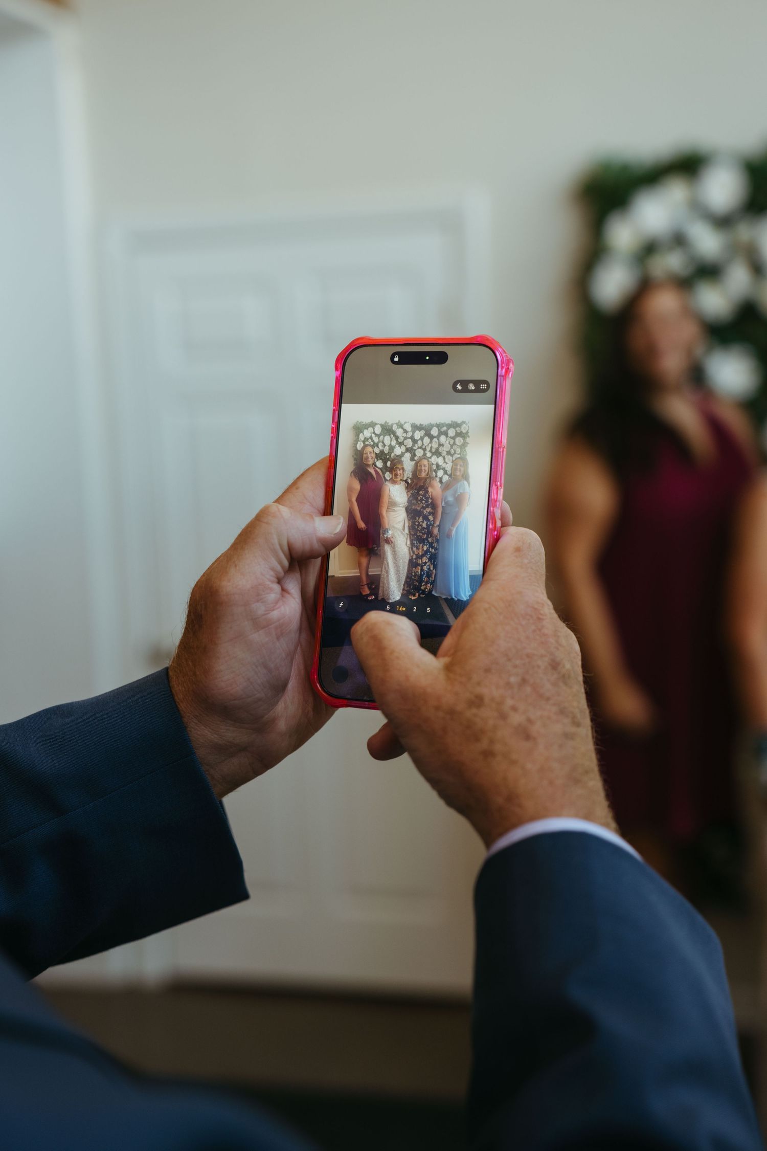 Person in suit photographing women at wedding venue using smartphone with red case, floral arch visible in background.