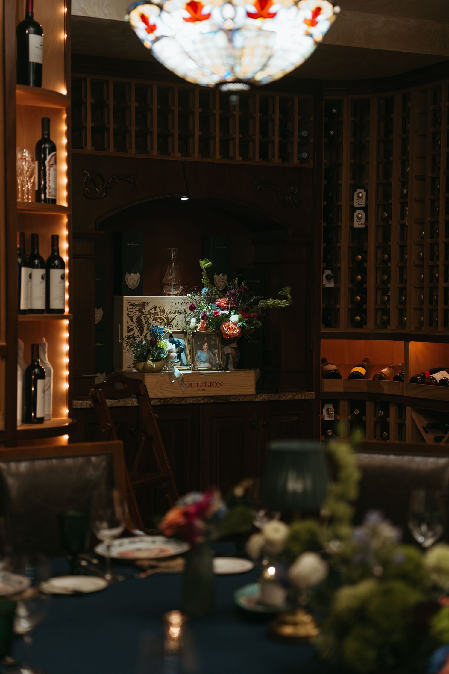 Elegant wine cellar dining room with floral centerpiece, chandelier, and floor-to-ceiling wine racks in warm lighting.
