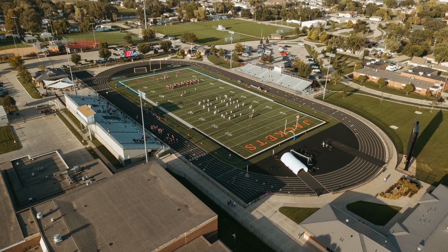 Aerial view of a high school football field with running track, surrounded by buildings and trees in a small town.