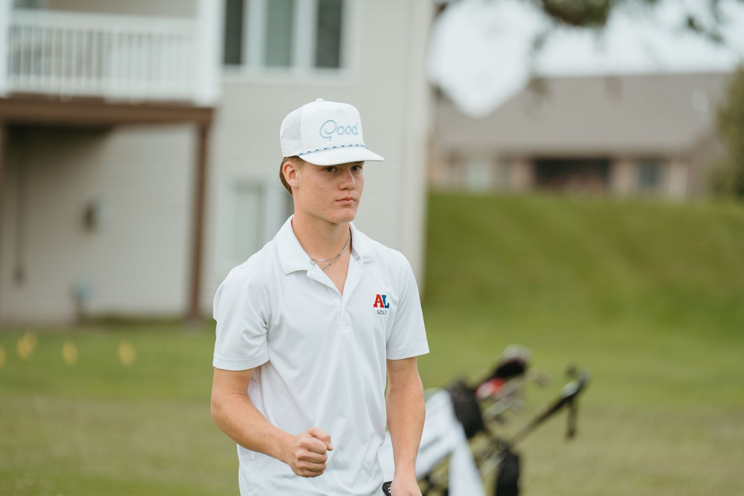 Young male golfer in white polo and cap standing on a golf course near a golf bag.